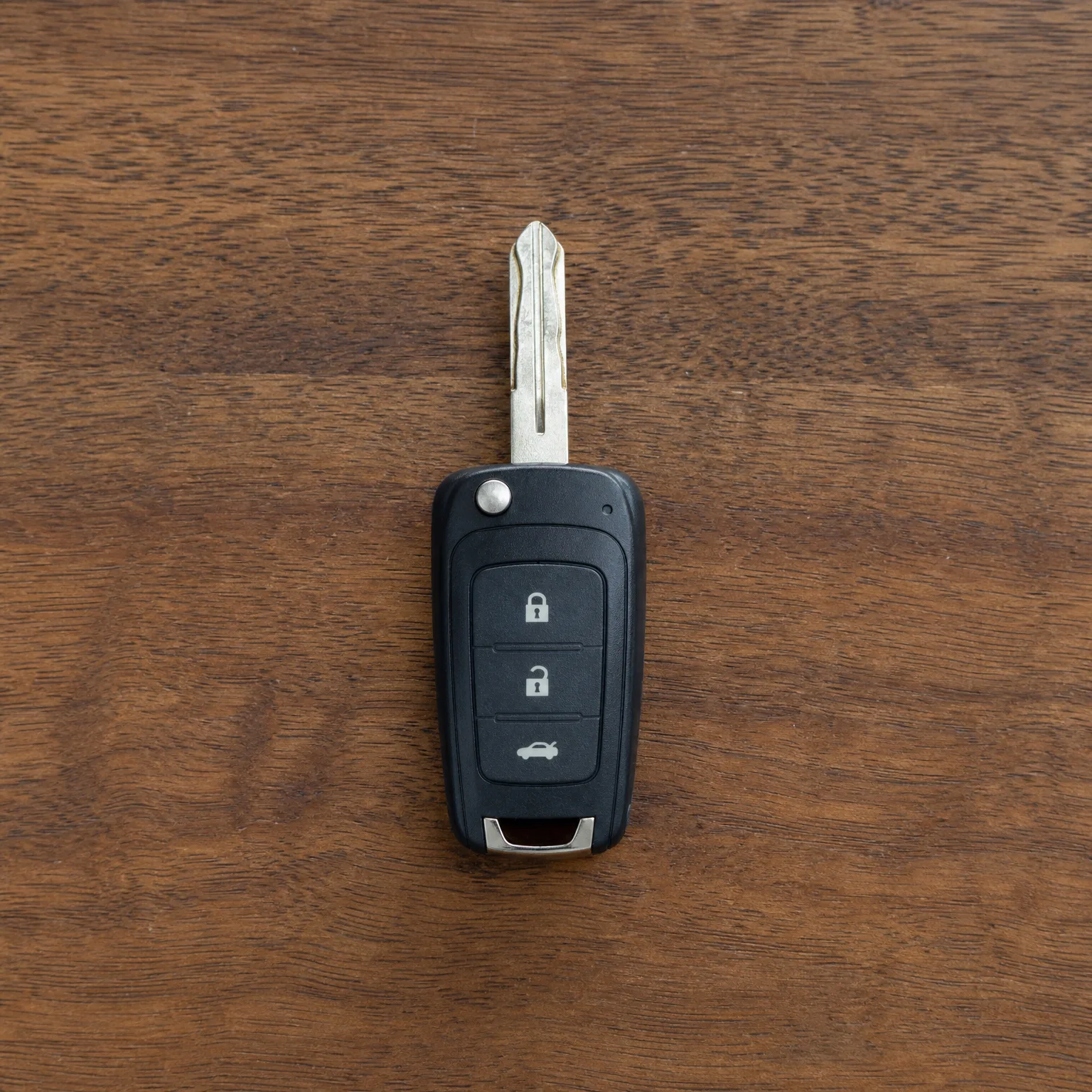 A black flip-style car key with three buttons (lock, unlock, trunk) and an extended metal blade, lying on a brown wood surface.