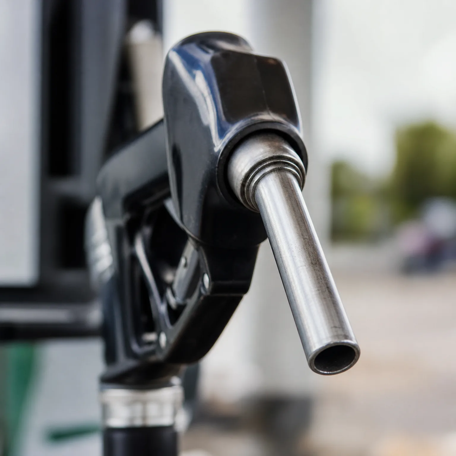Close-up of a black fuel nozzle with a metal spout mounted on a pump, with blurred greenery and pavement in the background.