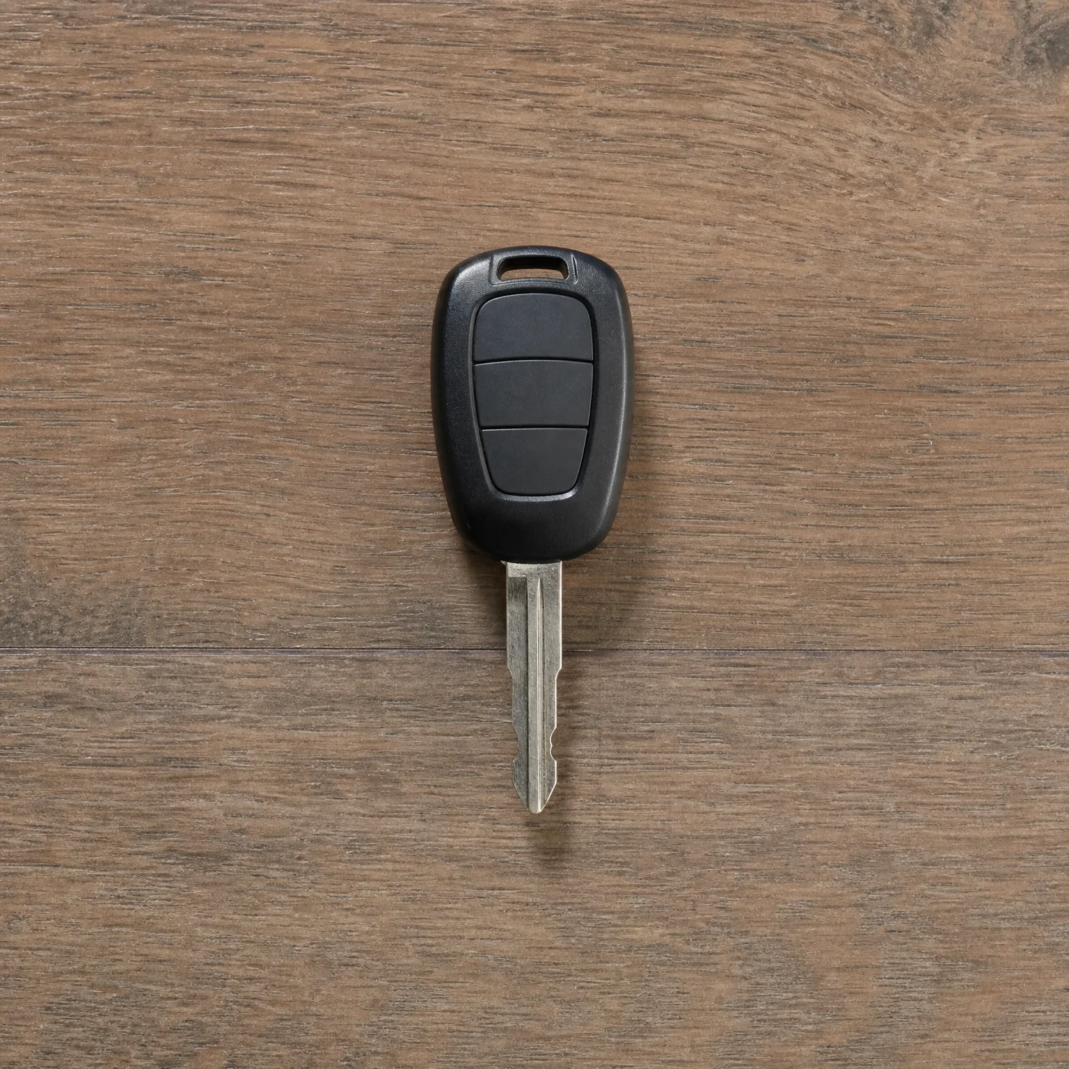 A black car key with a three-button remote head and uncut metal blade lying on a brown wood-grain surface.