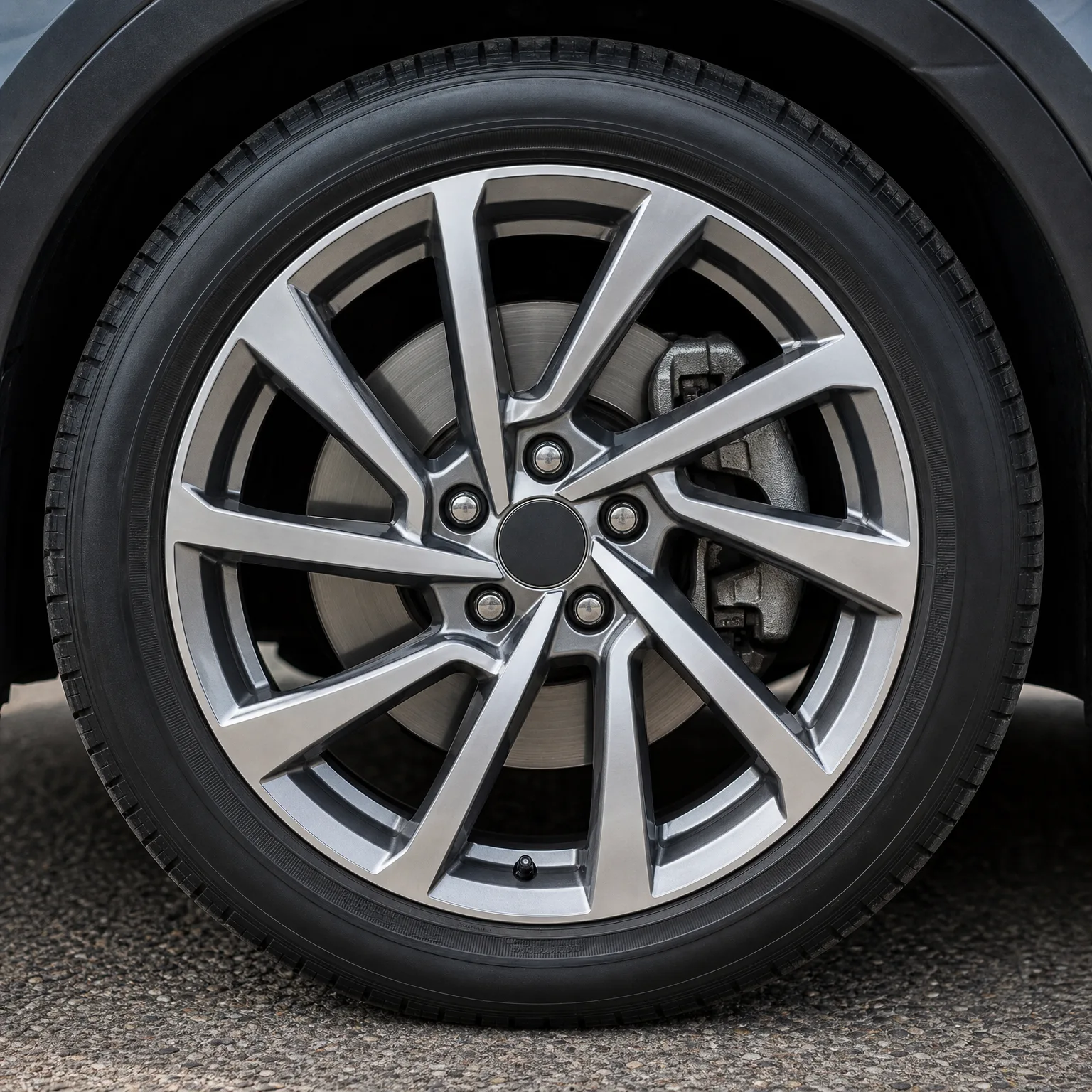 Close-up of a car's multi-spoke alloy wheel and tire, showing the brake disc and caliper behind the spokes, with asphalt pavement below.