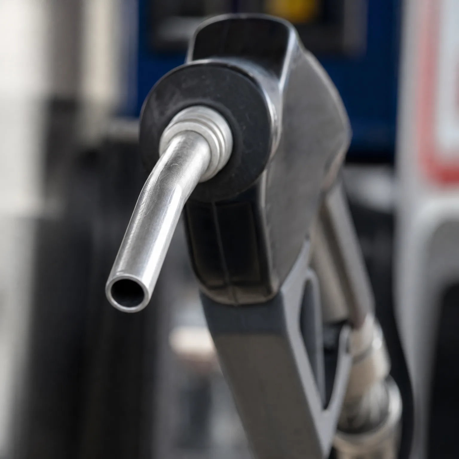 Close-up of a metal fuel pump nozzle with its spout pointing left, against a blurred background.