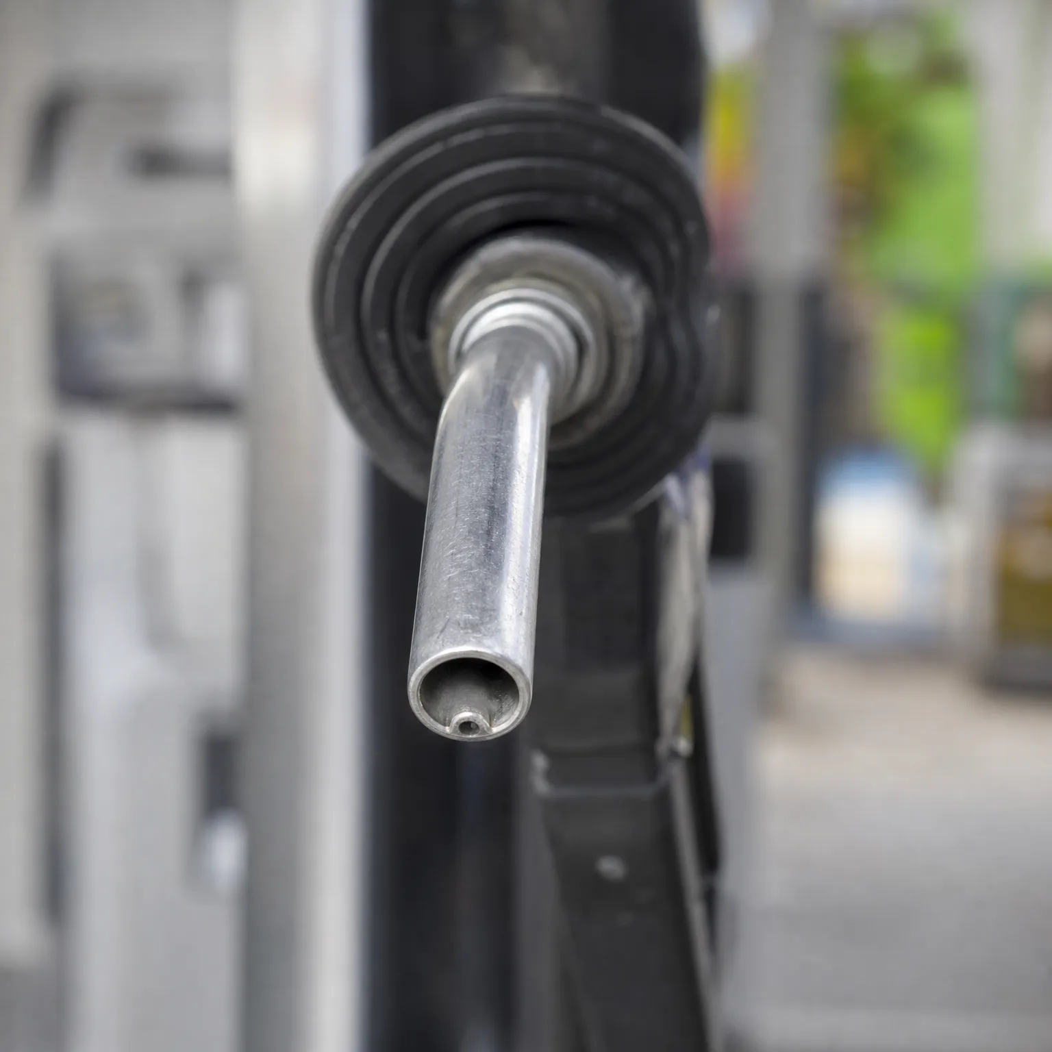 Close-up of a metal fuel nozzle spout with black rubber vapor-recovery bellows, with a blurred background of green foliage and structures.