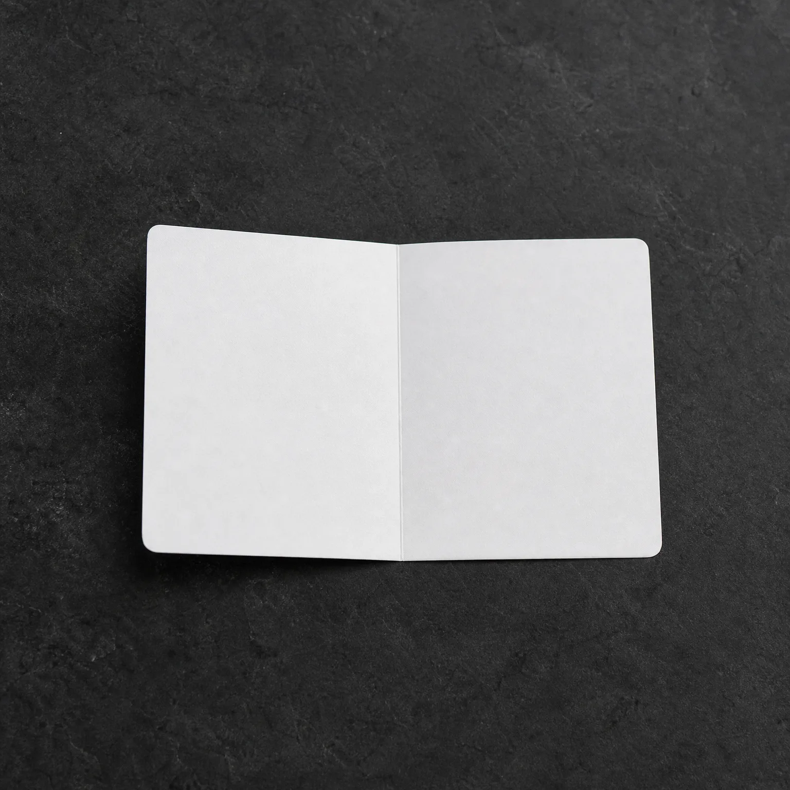 An open blank white folded card or booklet lying face up on a dark gray textured surface.