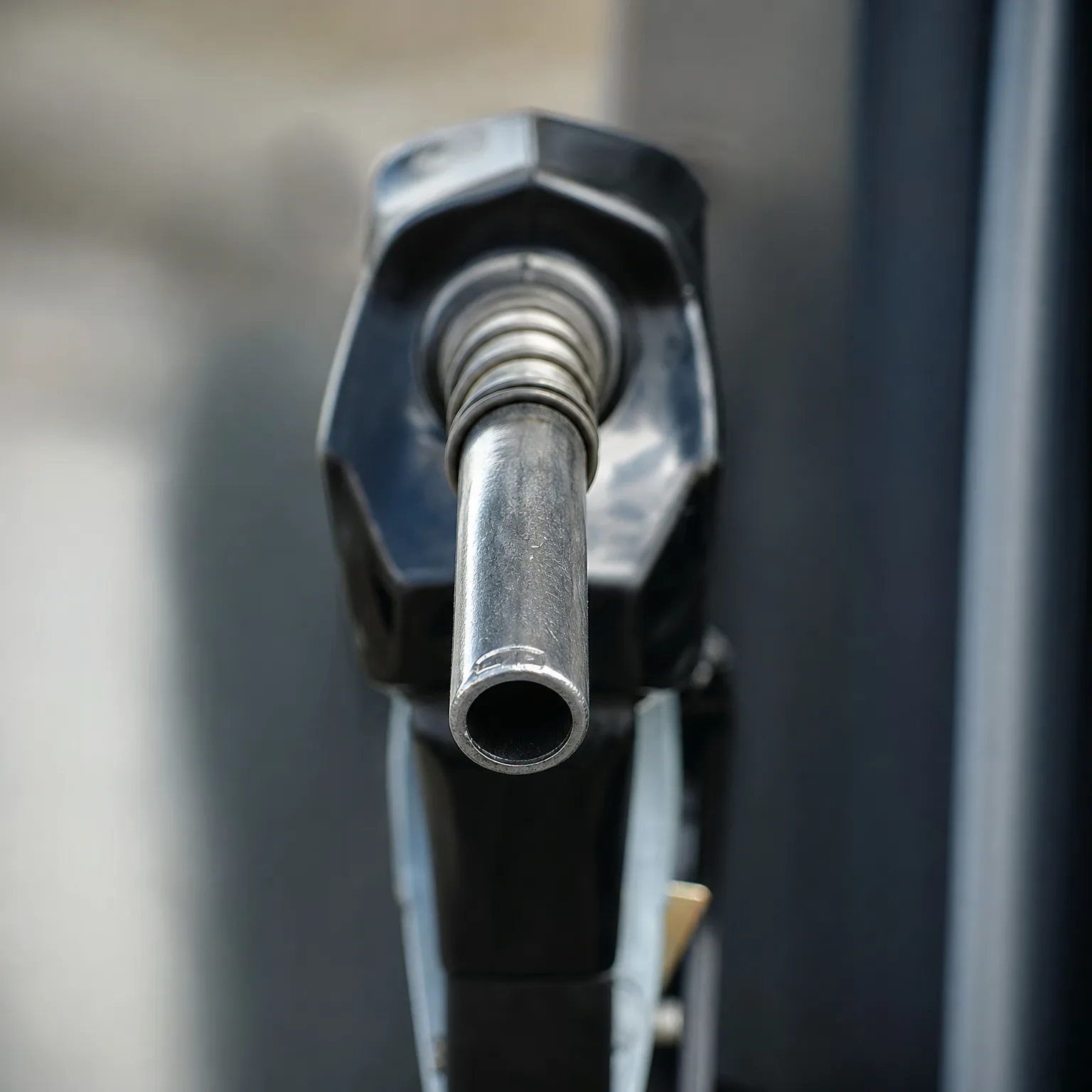Close-up of a black fuel nozzle with a metal spout, pointed toward the camera against a blurred gray background.