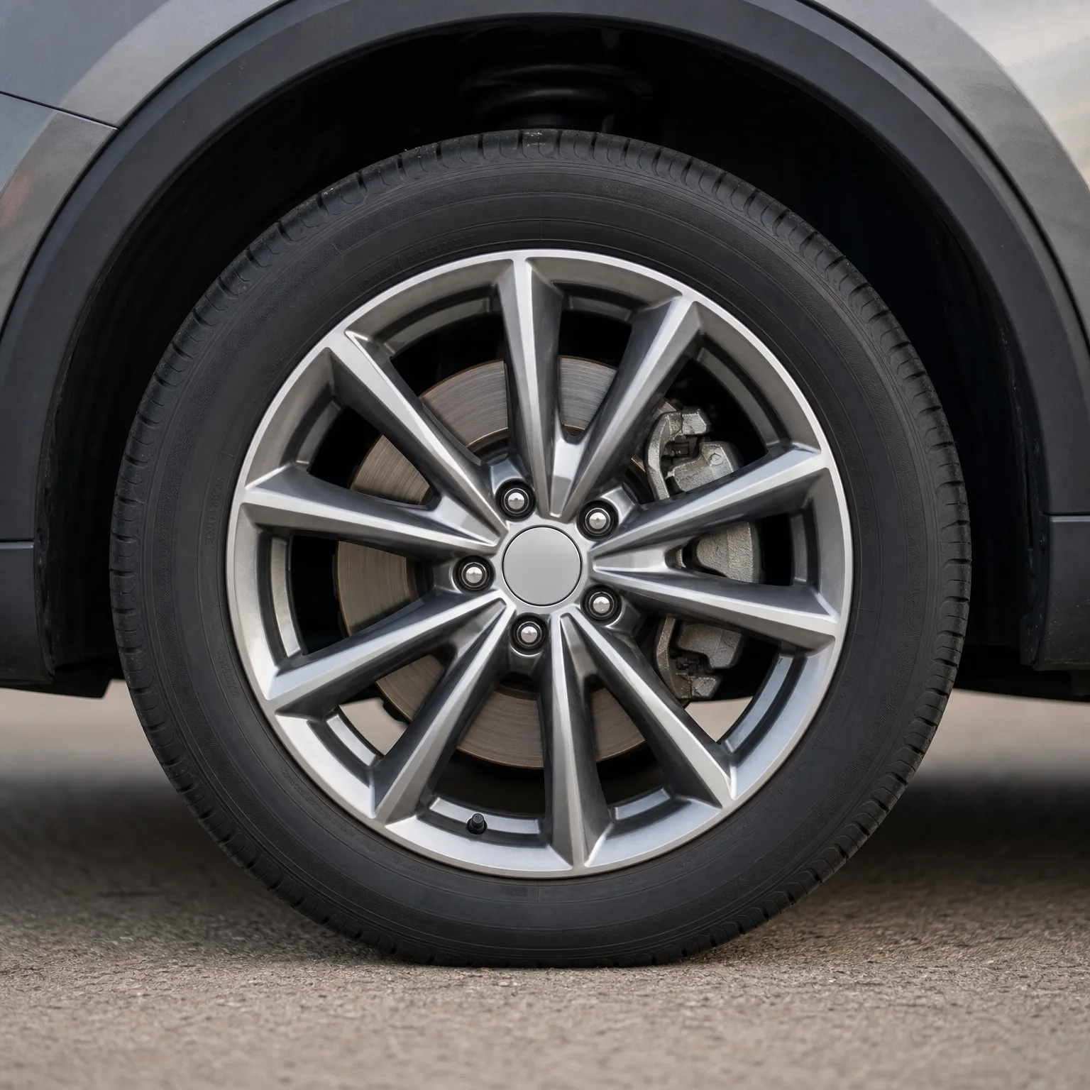 Close-up of a vehicle's front wheel with a multi-spoke silver alloy rim, black tire, and visible brake disc and caliper, parked on pavement.