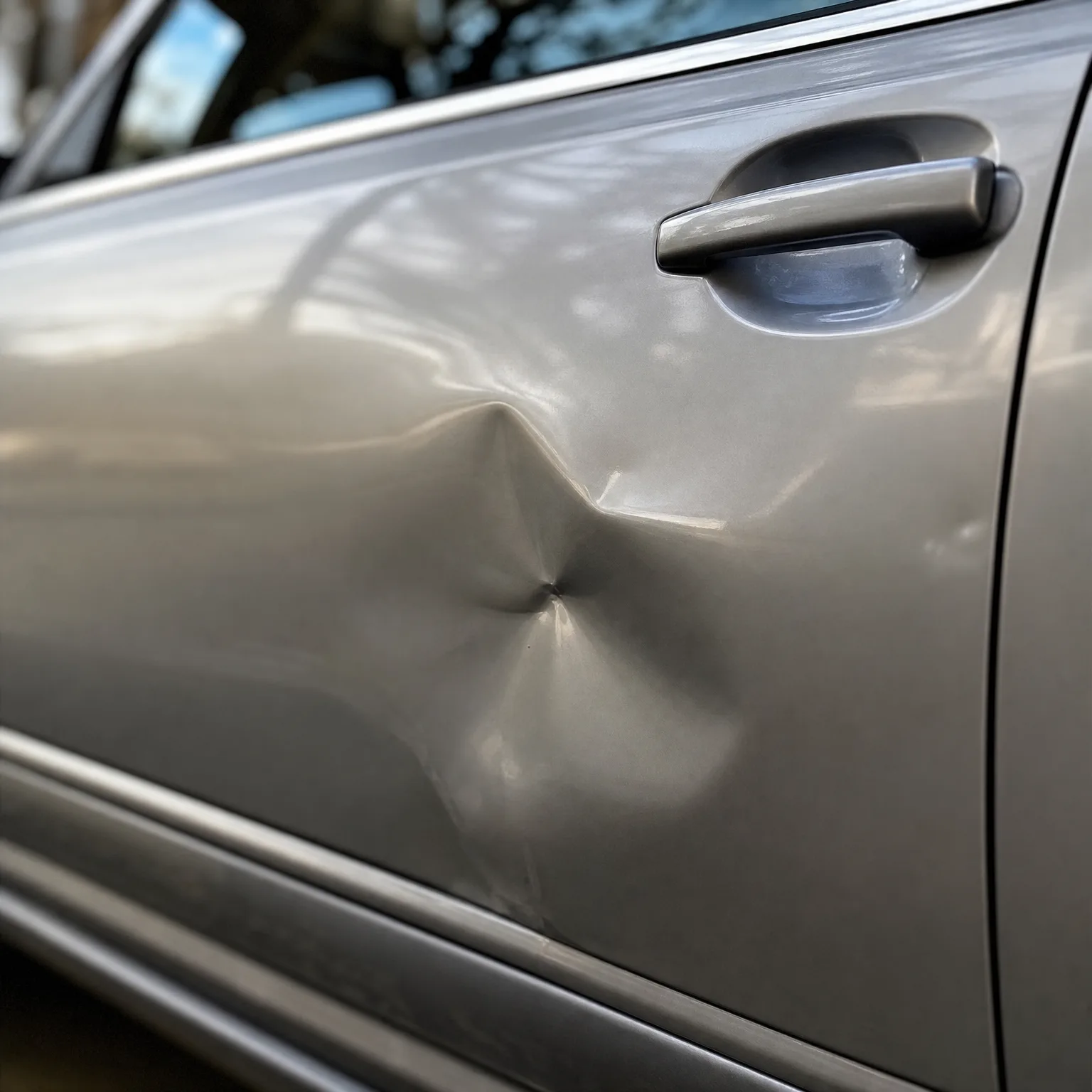 Close-up of a silver car's front door showing a large creased dent below the door handle, with blurred trees reflected in the window above.