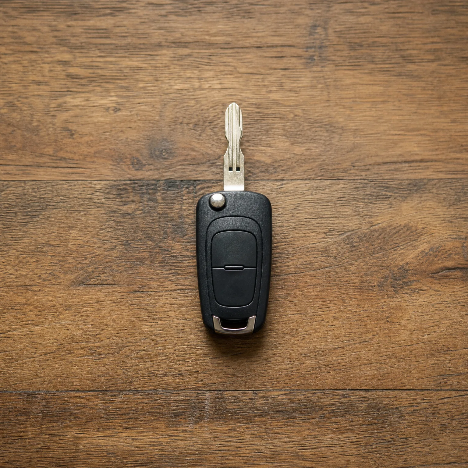 A black two-button flip car key with the metal blade extended, lying on a brown wooden plank surface.