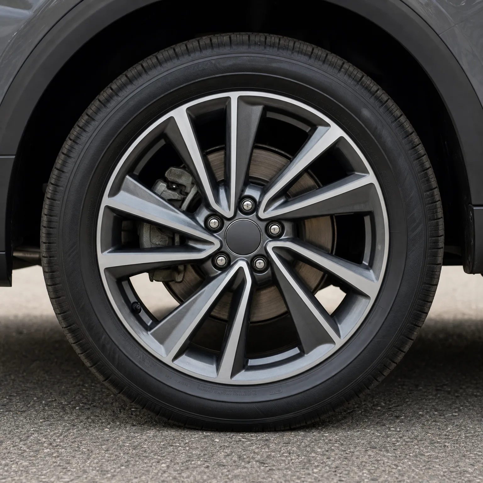 Close-up of a car's front wheel with a multi-spoke machined alloy rim and black tire, showing the brake disc and caliper behind it, parked on asphalt.