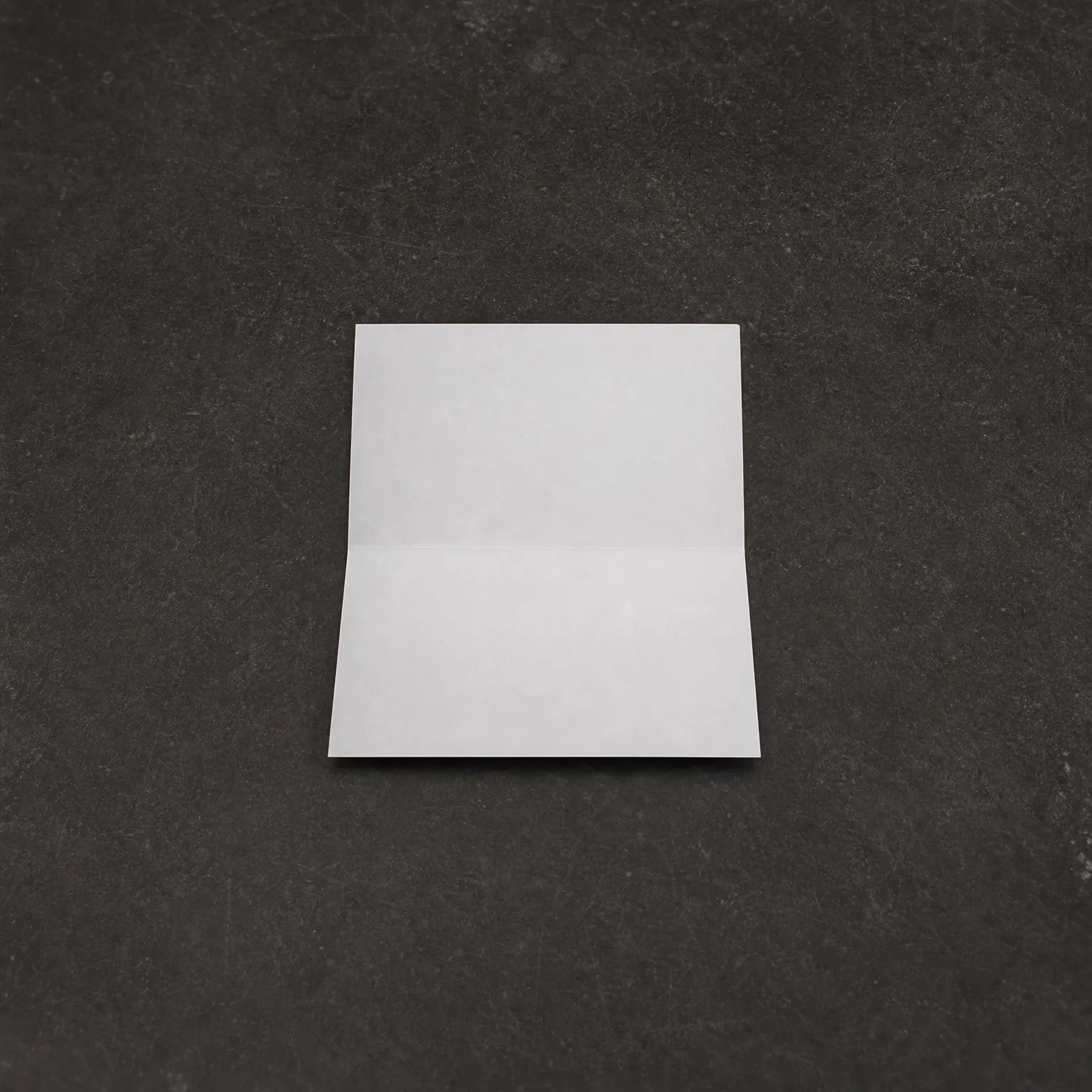 A blank white sheet of paper with a horizontal crease across its middle, lying on a dark gray textured surface.