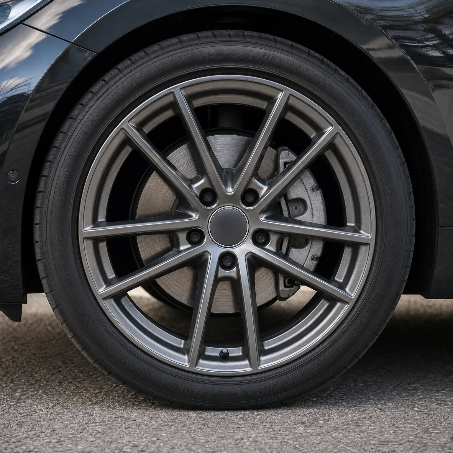 Close-up of a dark-painted car's front wheel with a gray multi-spoke alloy rim and low-profile tire, parked on asphalt.