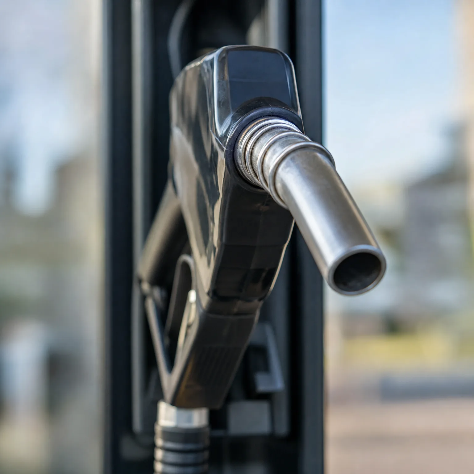 Close-up of a black fuel nozzle with a metal spout resting in a fuel pump, with a blurred outdoor background.