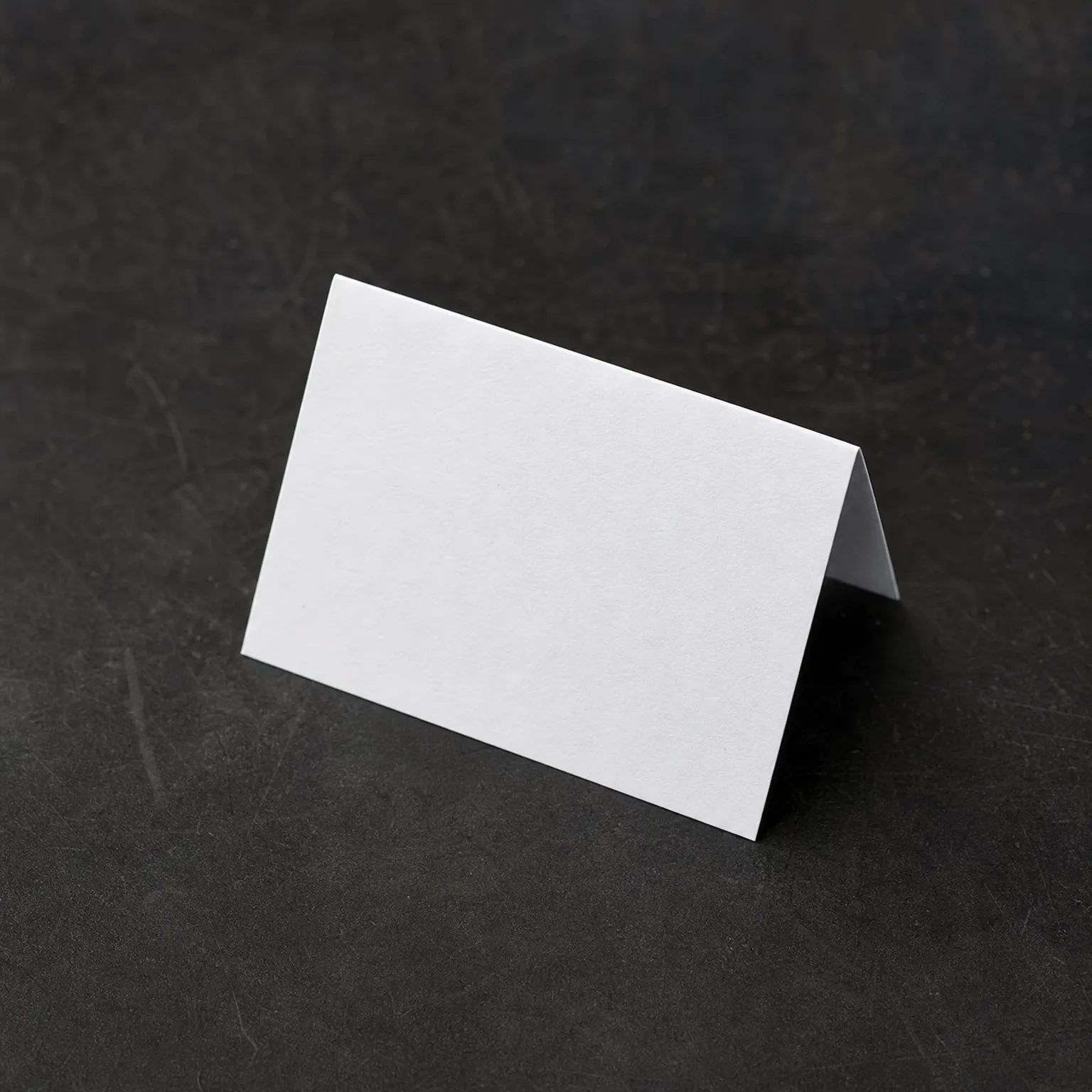 A blank white folded tent card standing on a dark gray textured surface.