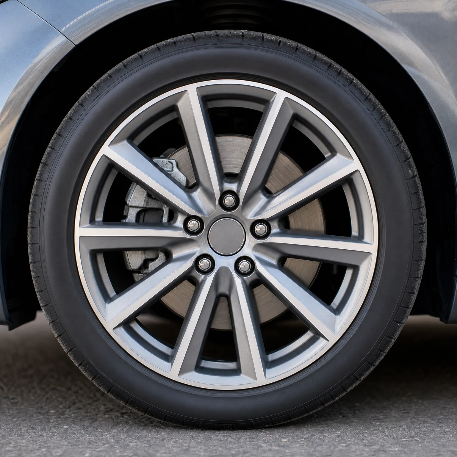 Close-up of a car's front wheel with a ten-spoke two-tone alloy rim, five lug nuts, visible brake disc and caliper, and low-profile tire, on pavement.