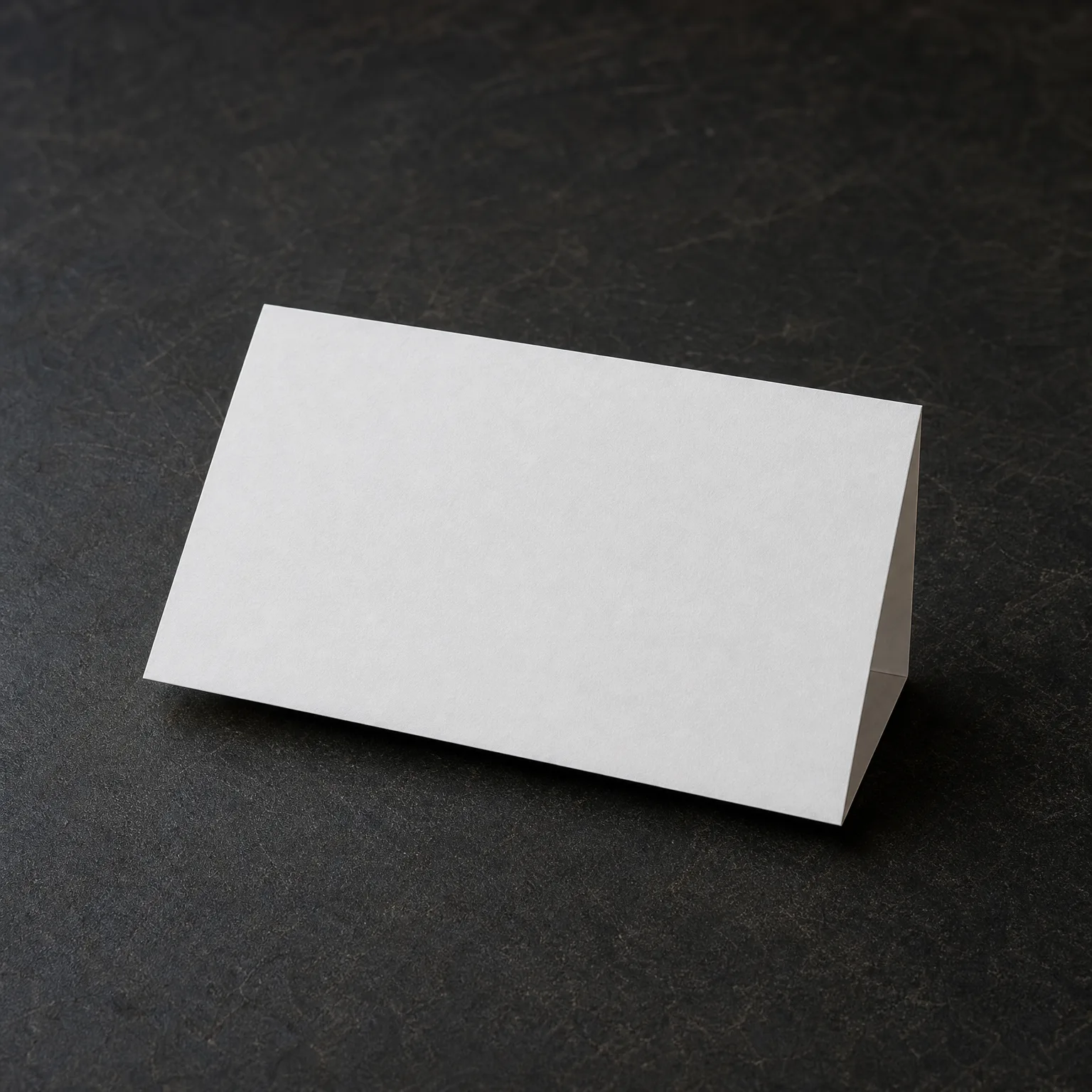 A blank white folded tent card standing on a dark textured gray surface.