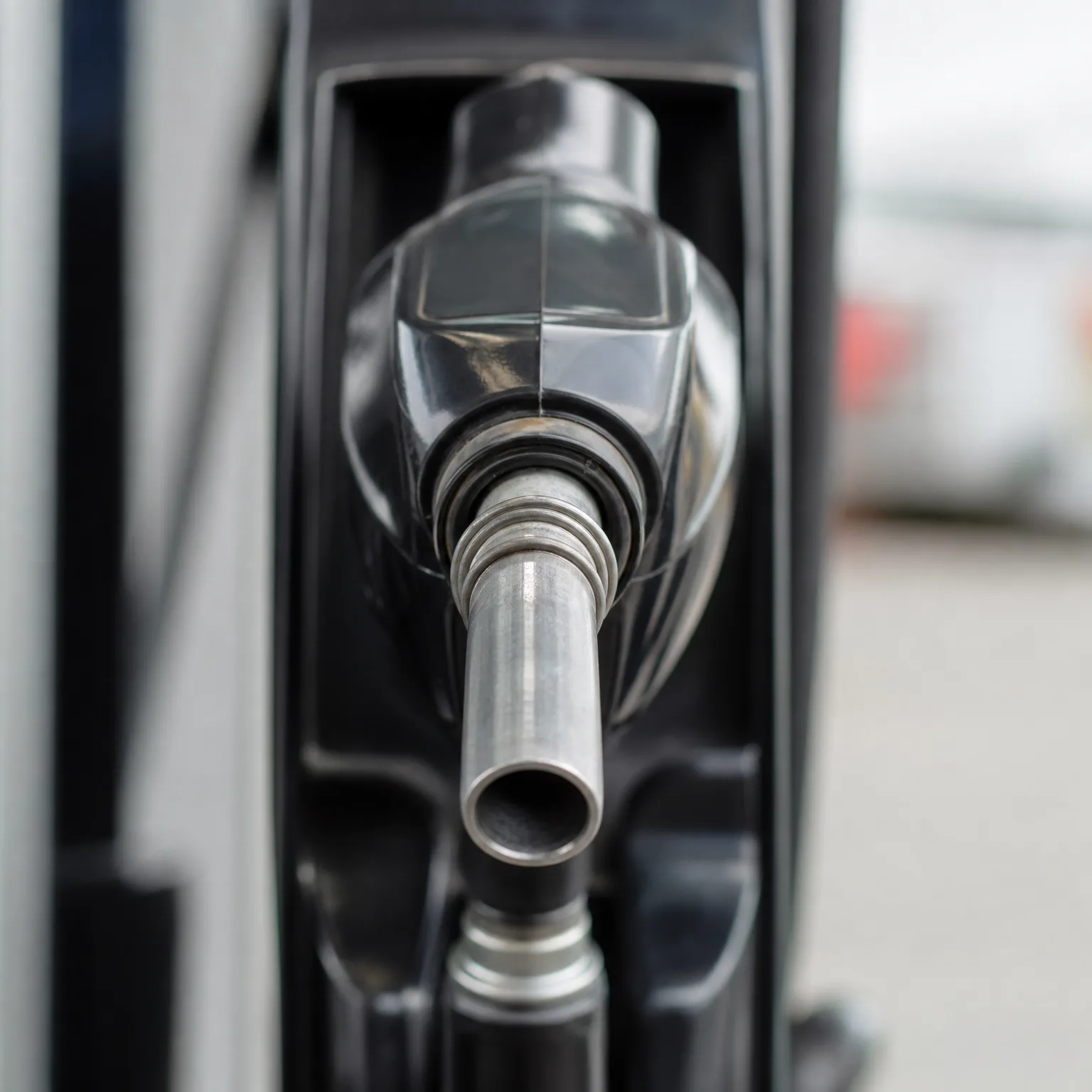 Close-up of a metal fuel nozzle resting in the holster of a dark-colored fuel dispenser, with a blurred paved background.