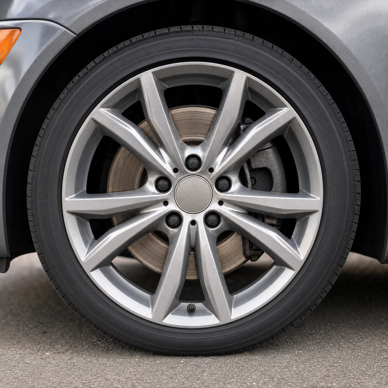 Close-up of a silver multi-spoke alloy wheel with a low-profile tire on a gray car, with the brake disc and caliper visible behind the spokes, parked on asphalt.
