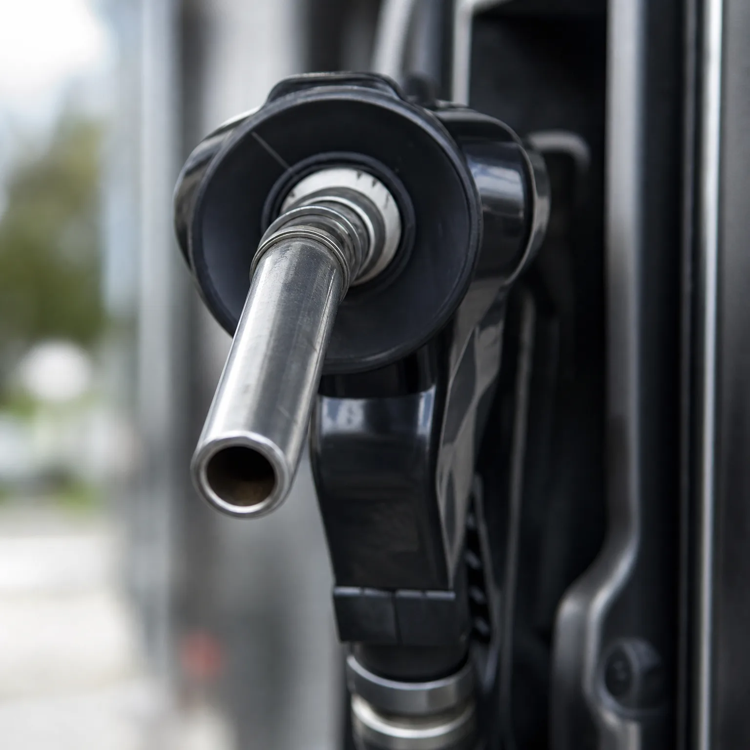 Close-up of a black fuel nozzle with a metal spout mounted on a pump, with blurred greenery in the background.