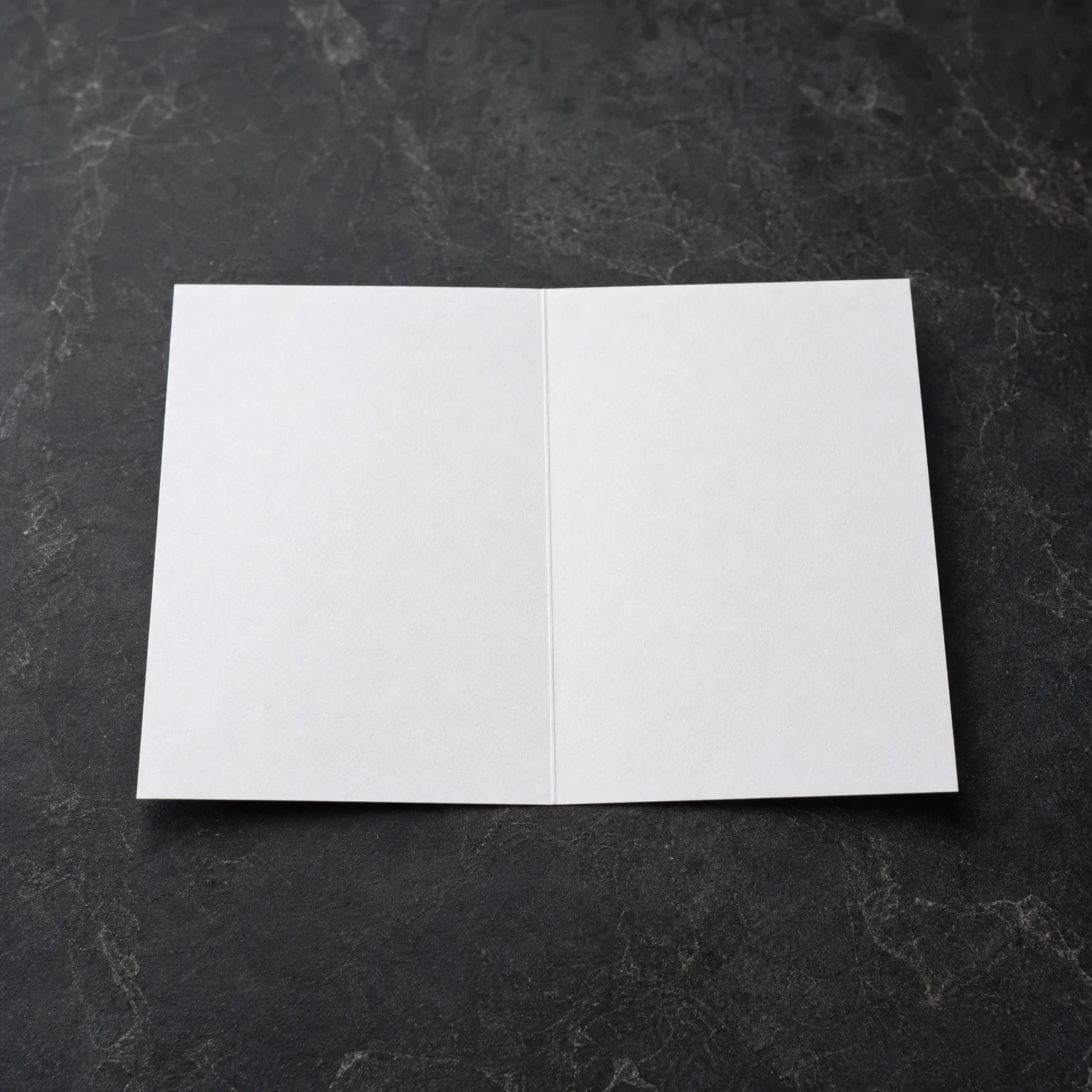 A blank white folded card lying open on a dark grey textured stone surface.