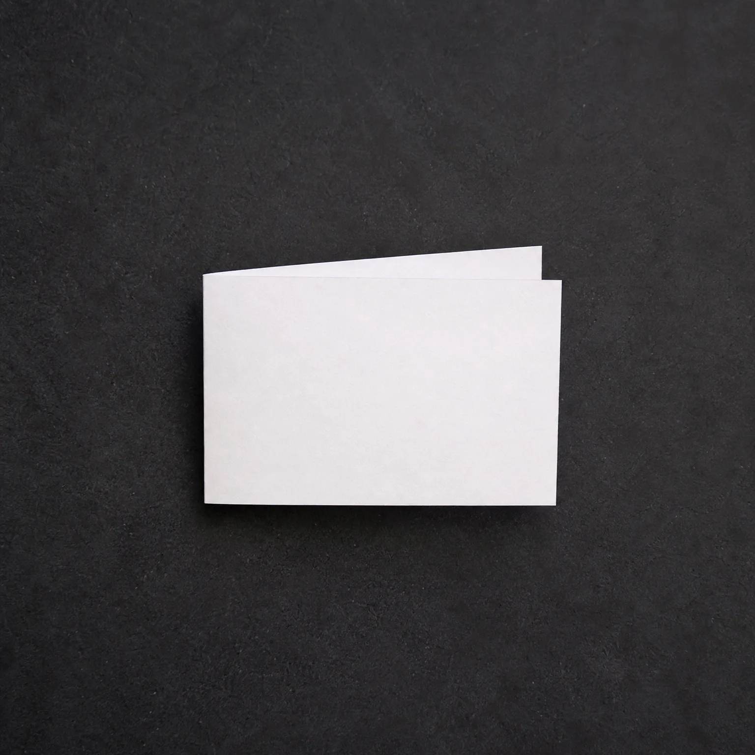 A blank white folded card lying on a dark gray textured surface, photographed from above.