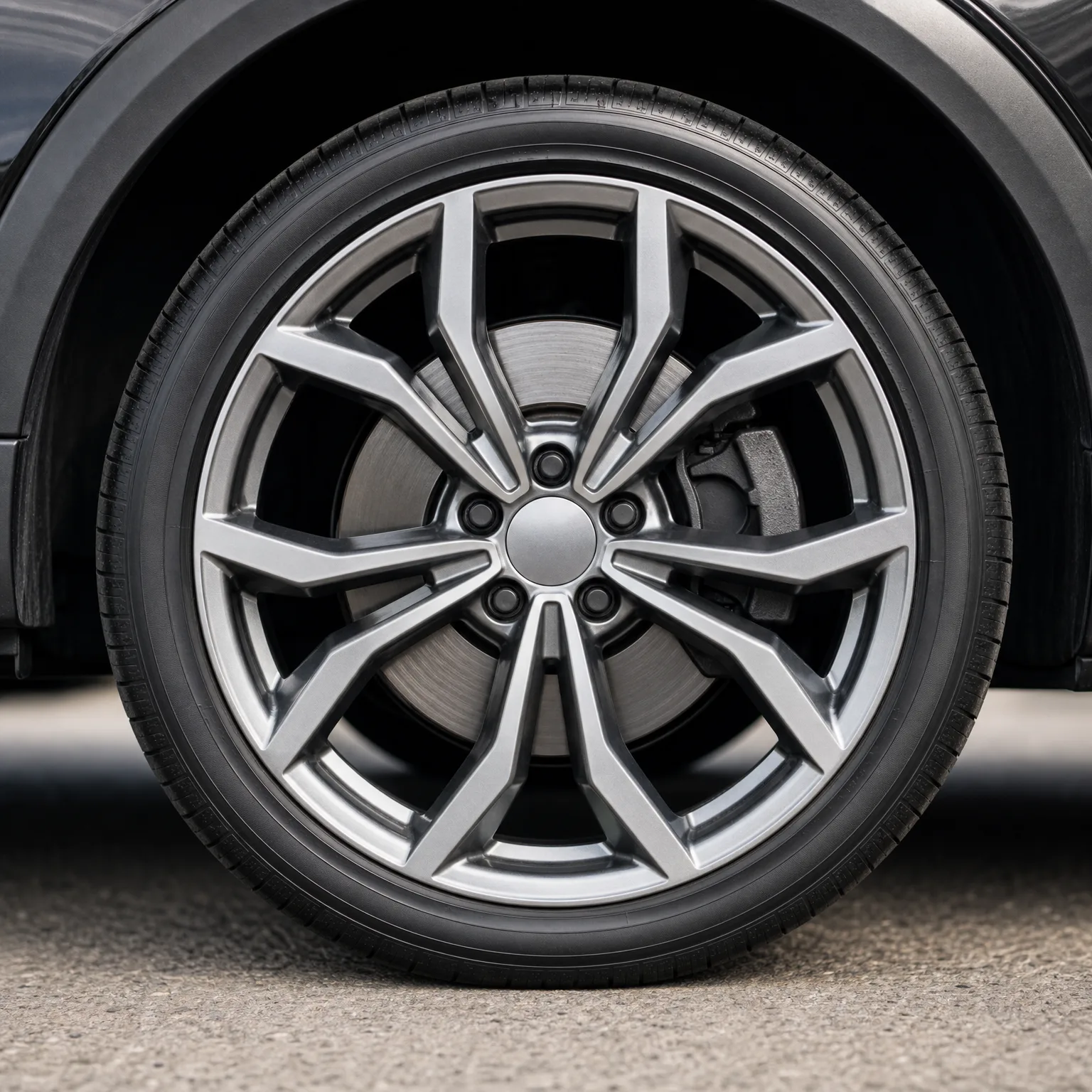 Close-up of a dark vehicle's front wheel with a multi-spoke silver alloy rim and low-profile tire, parked on pavement.