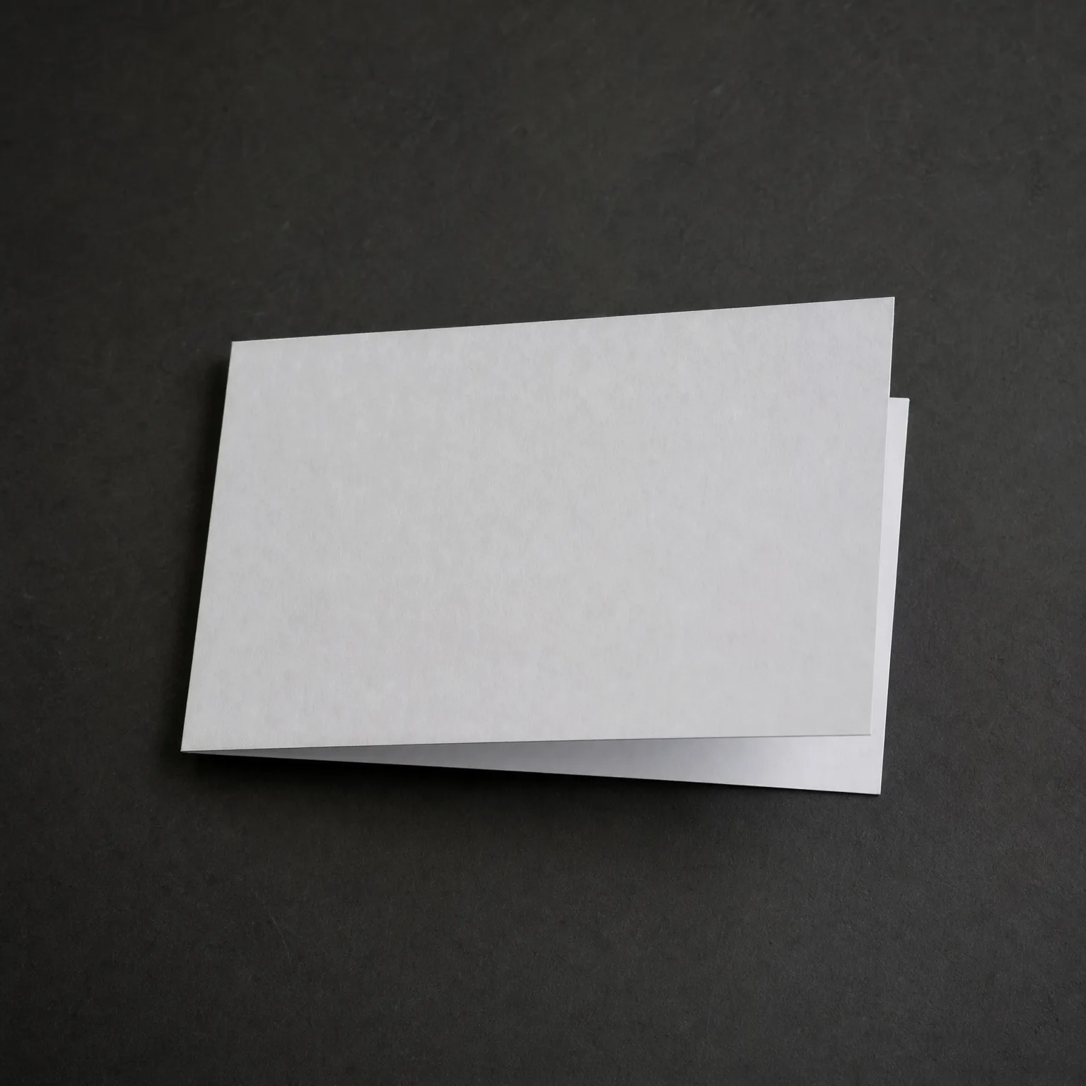 A blank white folded card lying on a dark gray textured surface, lit from above.