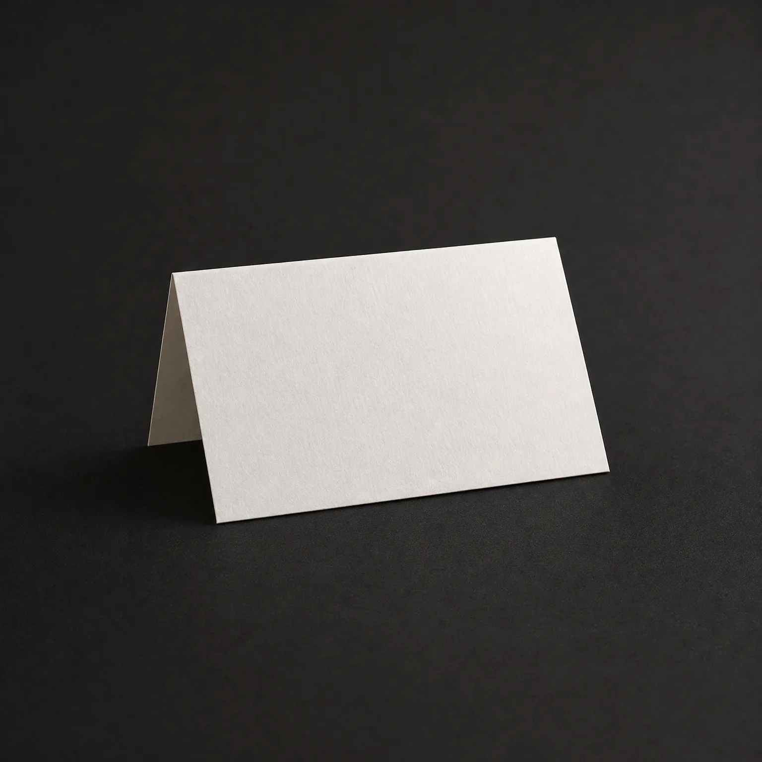 A blank white folded tent card standing upright on a dark gray surface against a dark background.