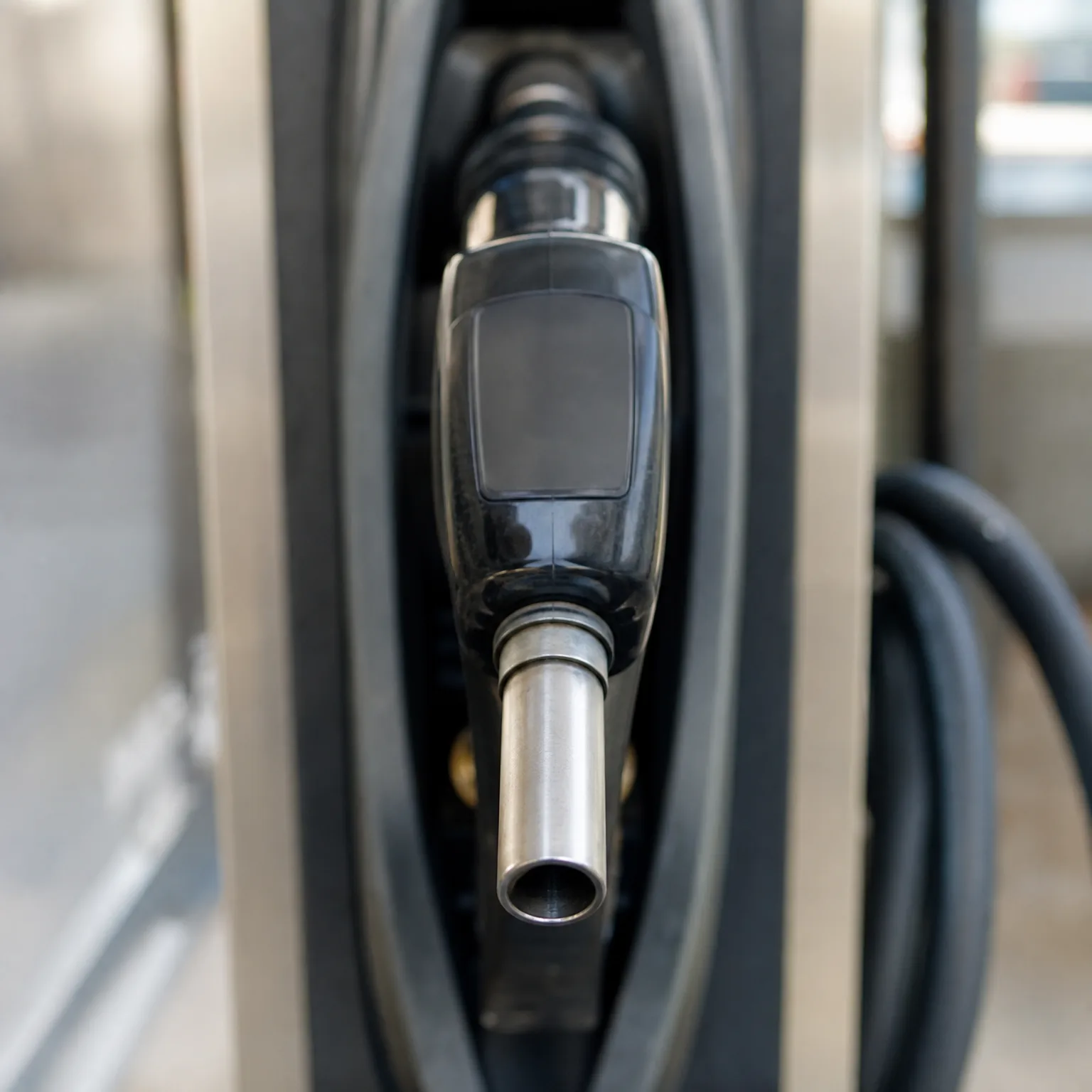 Close-up of a black fuel nozzle holstered in a gas pump, with a coiled hose visible to the right and a blurred background.
