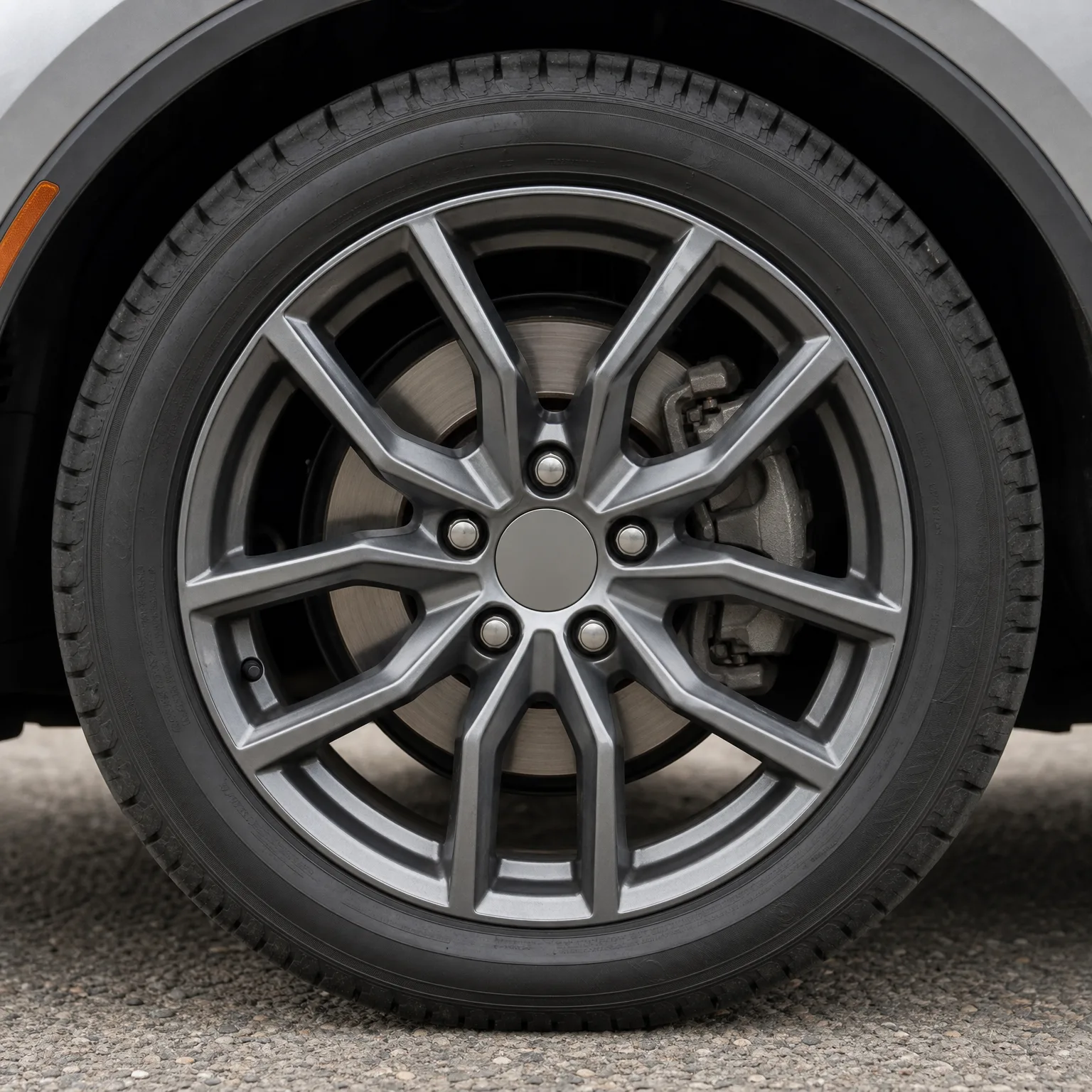Close-up of a car's gray multi-spoke alloy wheel and tire, with brake disc and caliper visible behind the spokes, over gravel pavement.