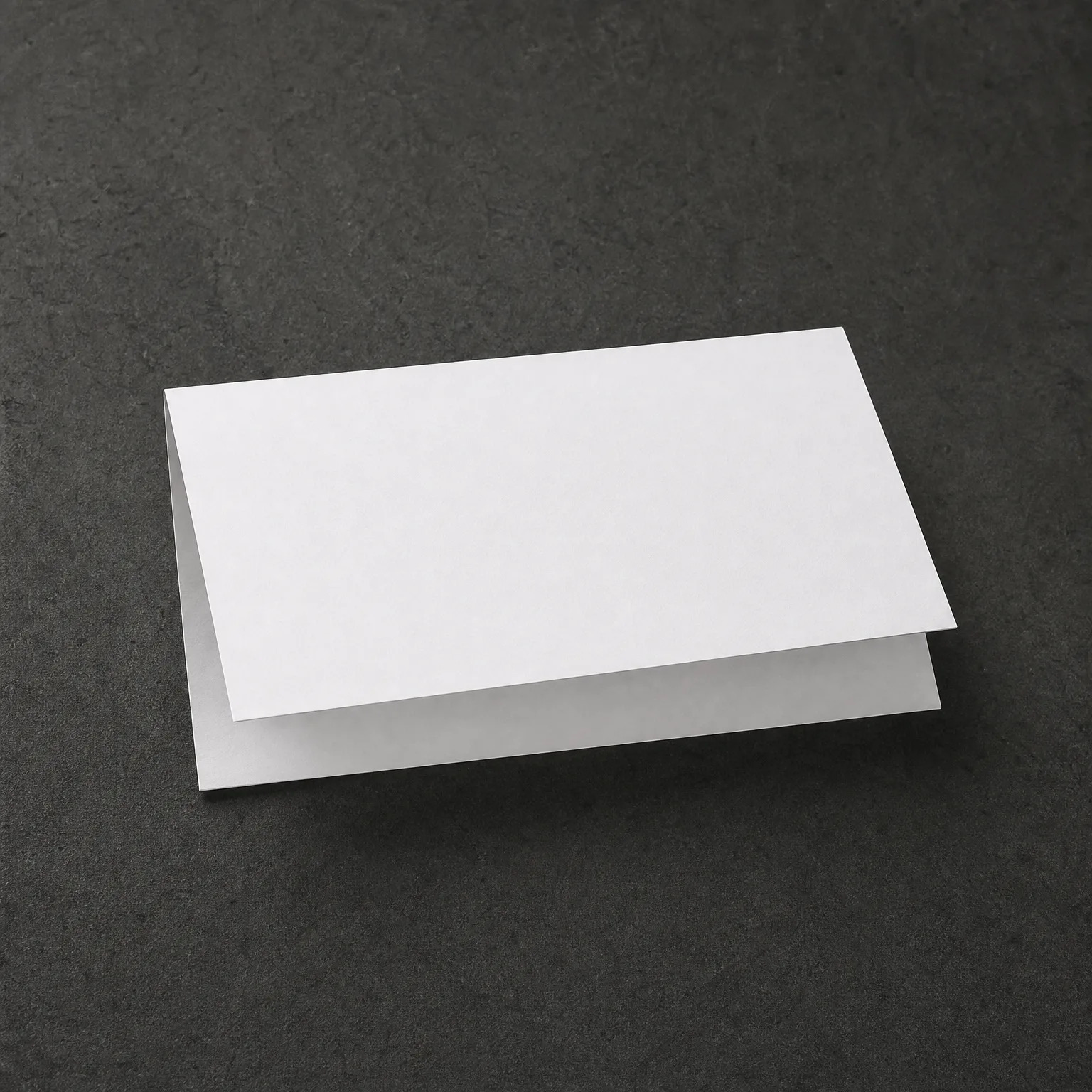 A blank white folded card stands tented on a dark gray textured surface.