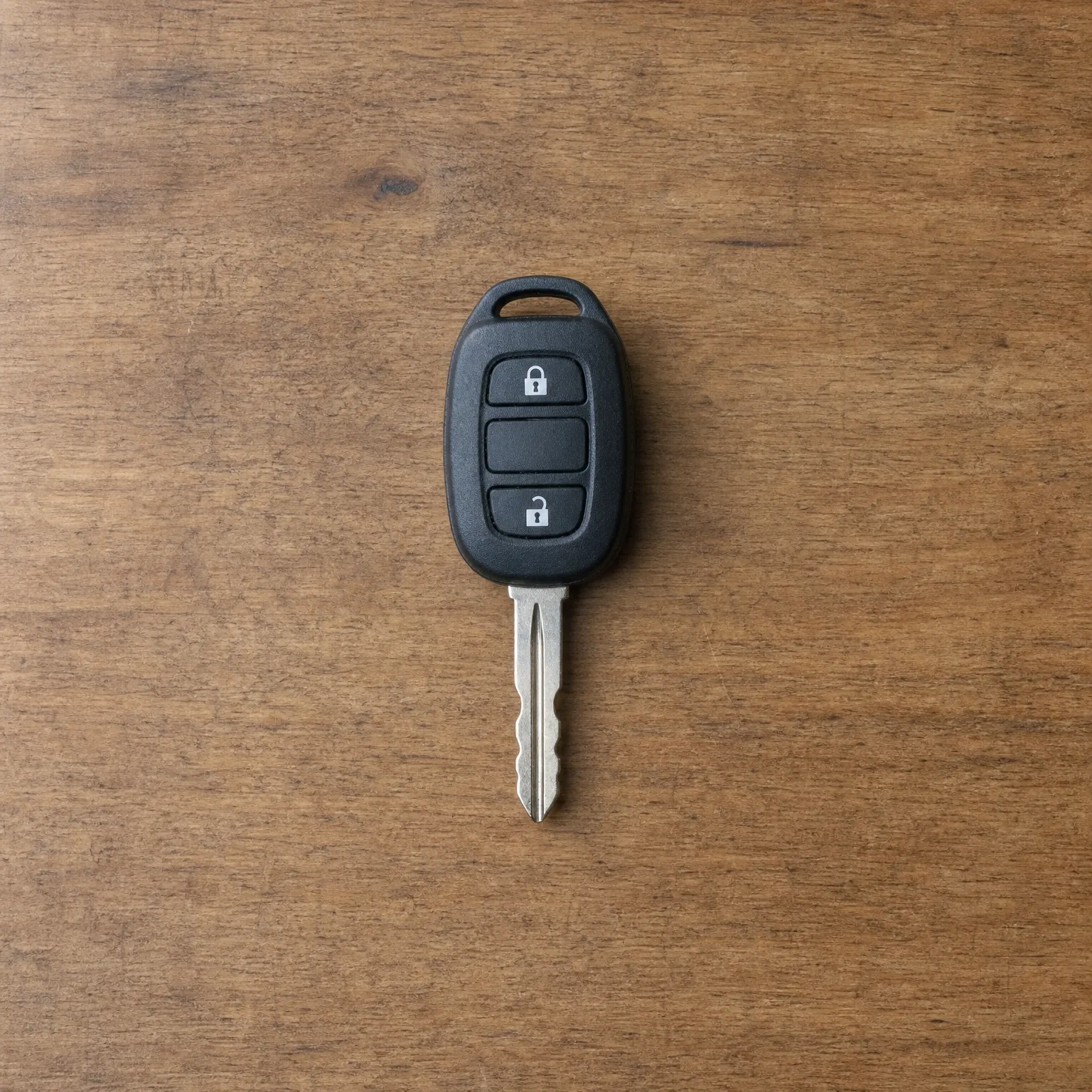 A black remote car key with three buttons, two showing lock and unlock padlock icons, and a cut metal blade, lying on a brown wooden surface.