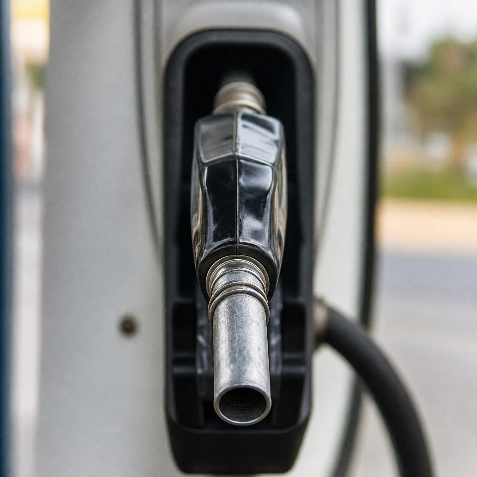 Close-up of a black fuel nozzle with a metal spout resting in its holster on a fuel dispenser, with a hose curving to the right and a blurred green and tan background.
