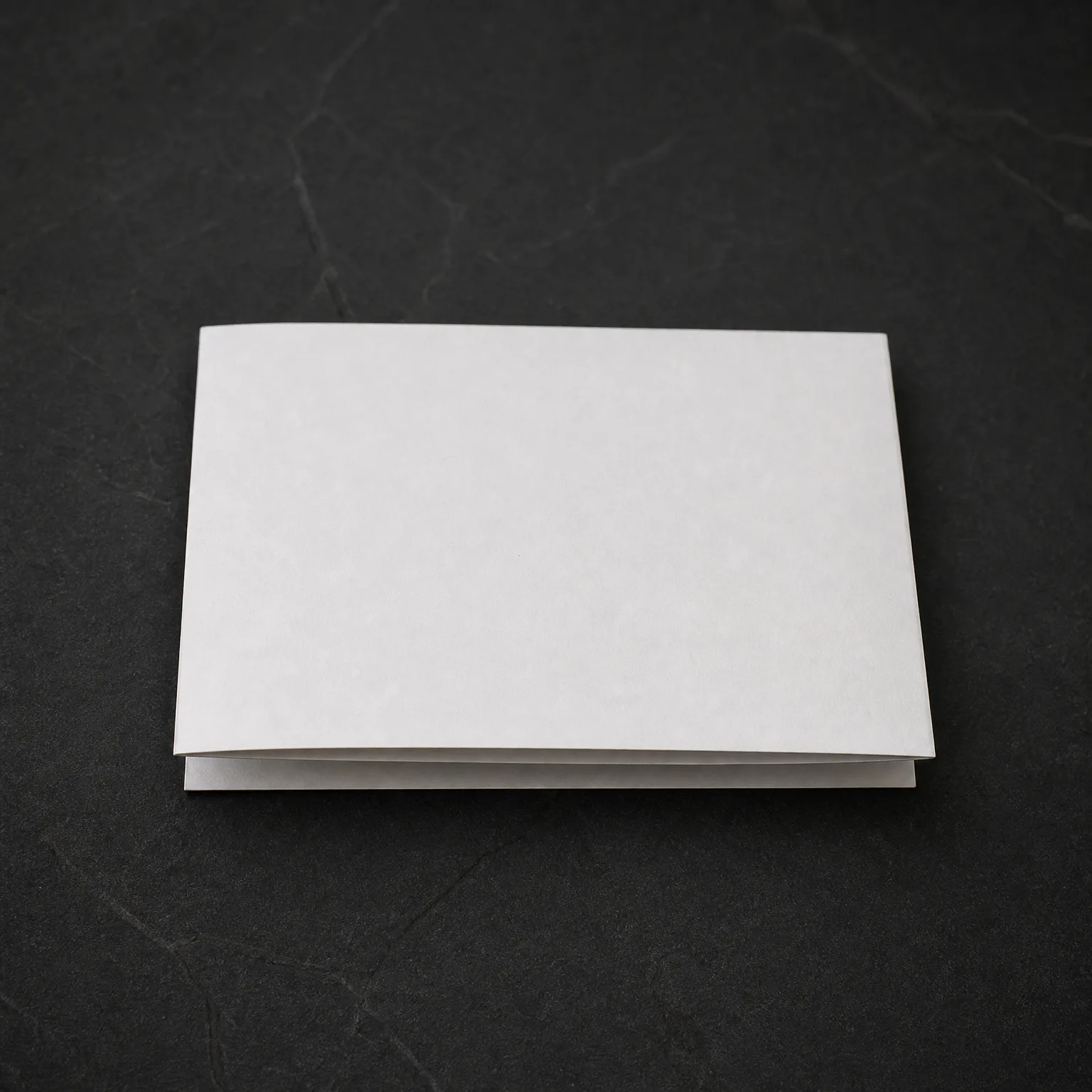 A blank white folded card lying closed on a dark gray textured surface.