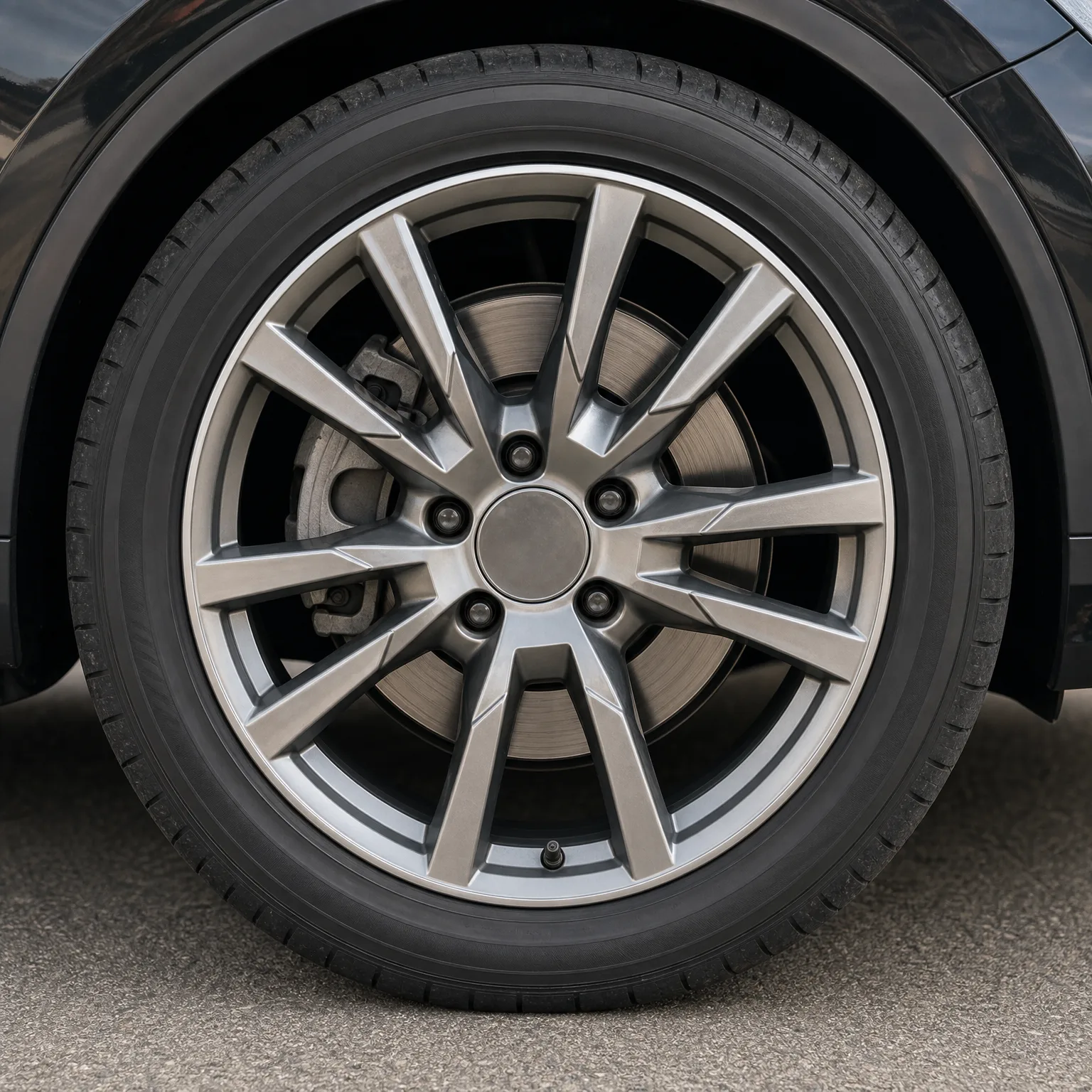 Close-up of a dark car's front wheel with a multi-spoke silver alloy rim, low-profile tire and visible brake disc and caliper, parked on gray pavement.