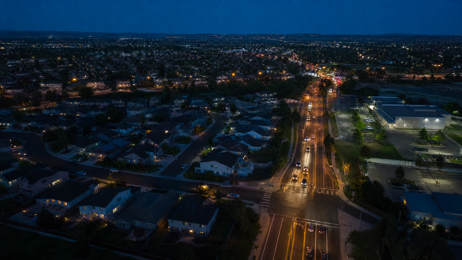 Aerial dusk view of a suburban city with a wide illuminated road cutting through residential neighborhoods and commercial buildings.