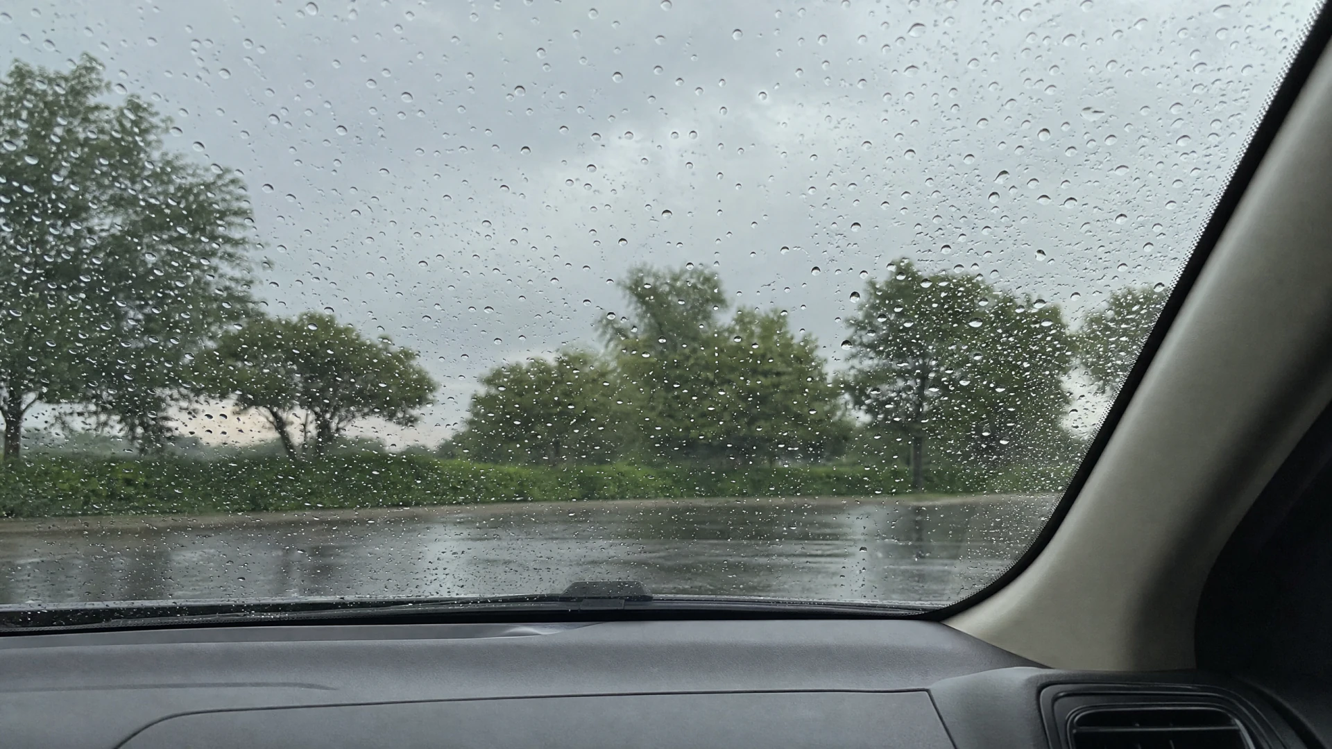 View from a car interior through a rain-droplet-covered windshield, showing a wet parking lot and green trees under a grey overcast sky.