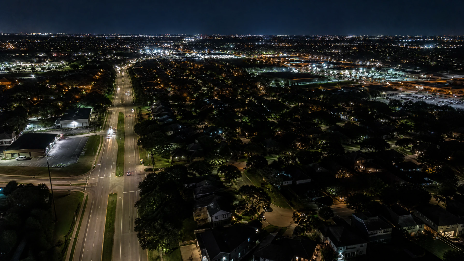 Aerial nighttime view of a suburban city with a wide road bisecting residential neighborhoods and lit commercial areas.