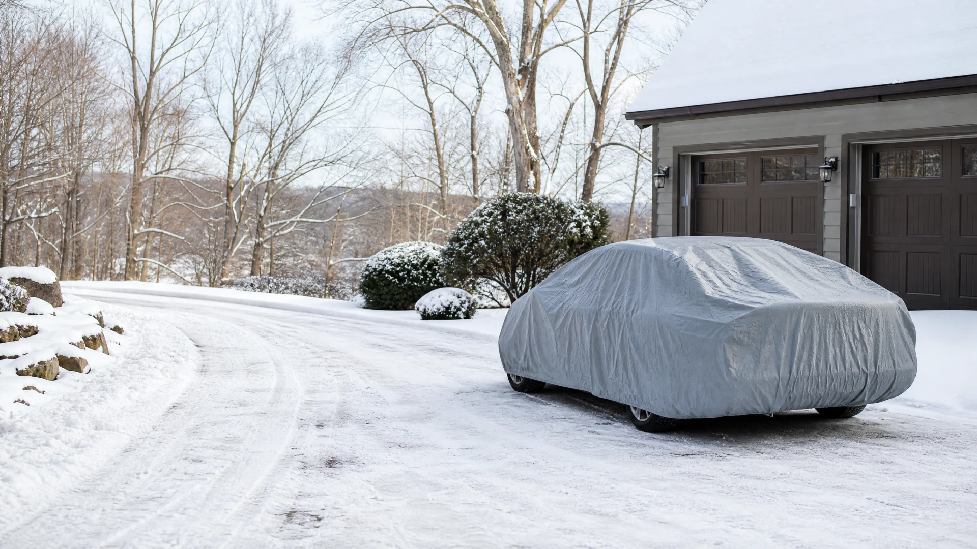 A gray car cover protects a vehicle parked in a snow-covered residential driveway beside a two-car garage.