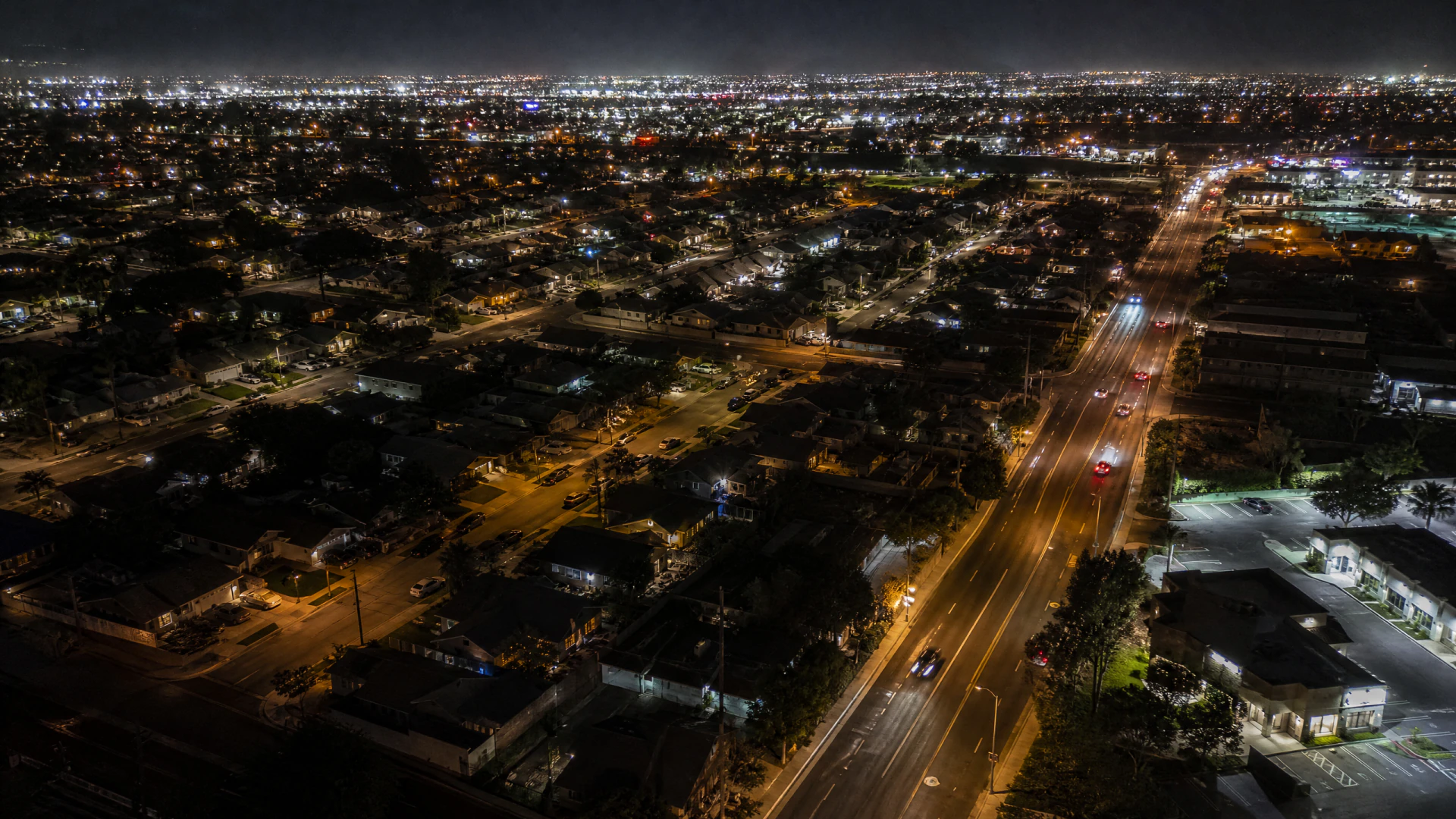 Aerial nighttime view of a sprawling urban neighborhood bisected by a wide illuminated multi-lane road.