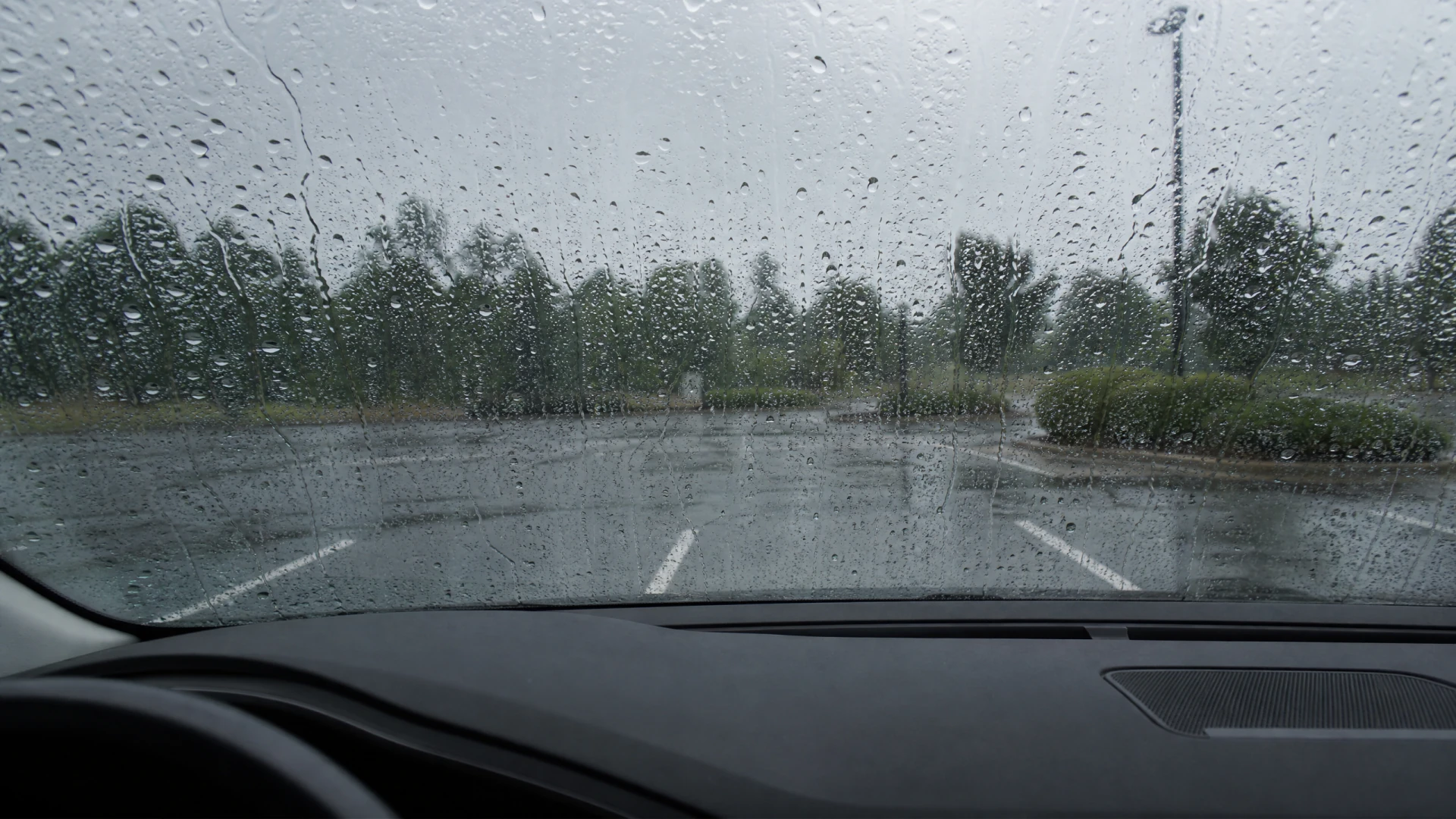 A rain-covered car windshield with a view of an empty wet parking lot surrounded by trees.