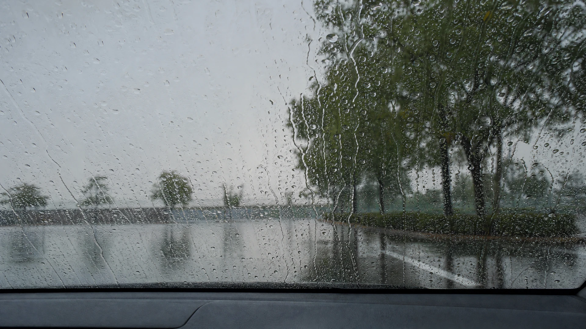 A rain-soaked car windshield with water droplets and wiper smears, through which a wet road and trees are visible under a grey sky.