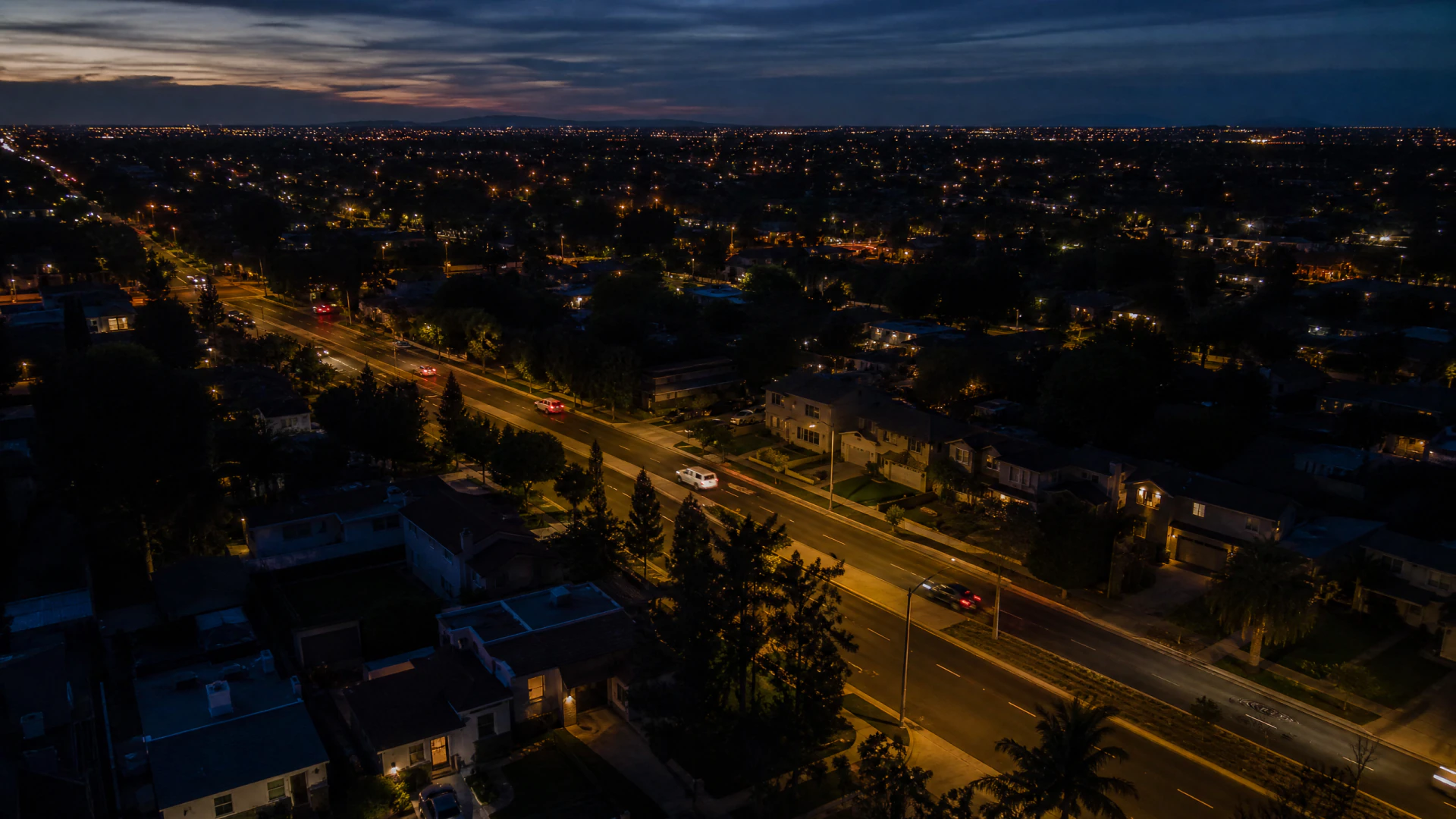 Aerial night photograph of a suburban boulevard lined with trees and orange street lights, surrounded by residential neighborhoods stretching to the horizon.
