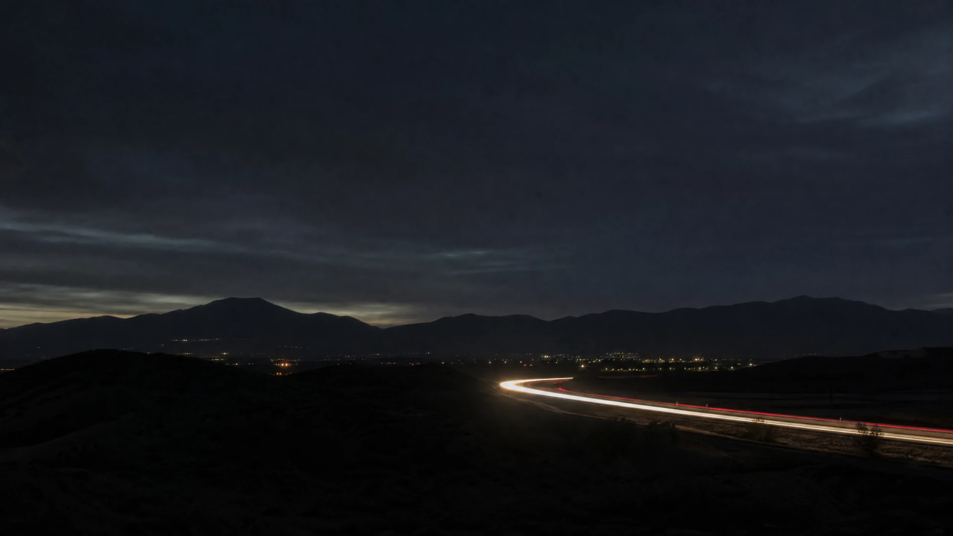Long-exposure night shot of white and red vehicle light trails curving along a road through a dark valley, with mountain silhouettes and distant city lights in the background.