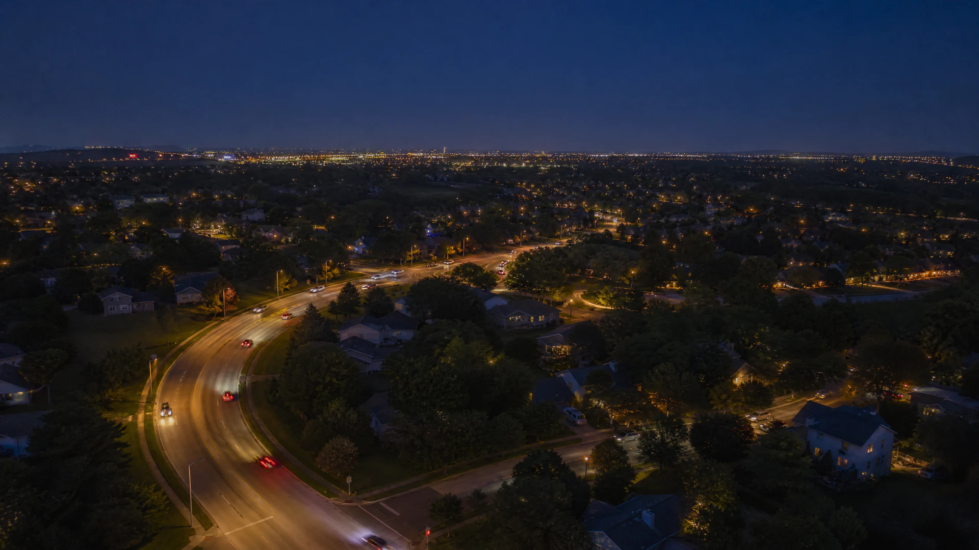Aerial night photograph of a curving suburban road with vehicle light trails, surrounded by tree-lined residential streets and a glowing city skyline in the distance.