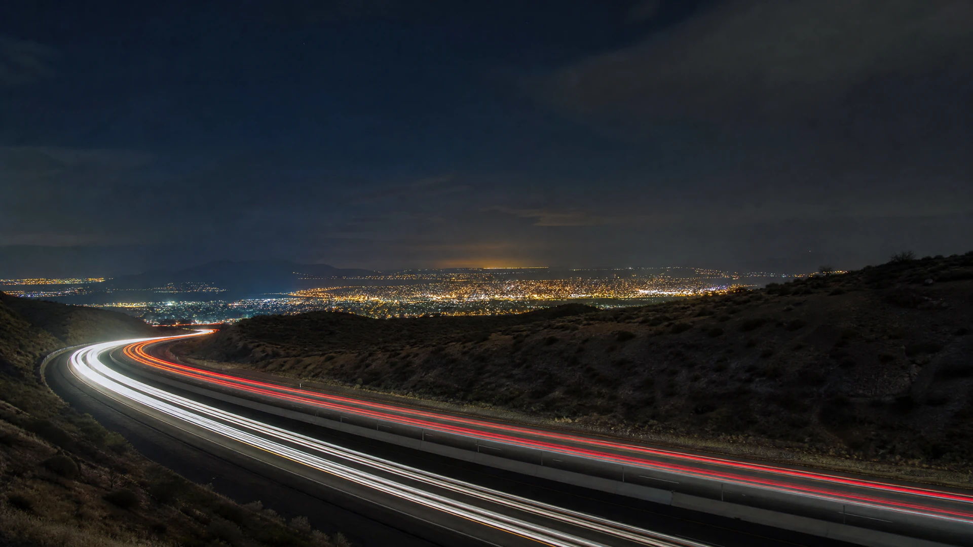 Long-exposure photograph of a curved highway at night, with white headlight and red taillight trails leading down toward a brightly lit city in a valley below.