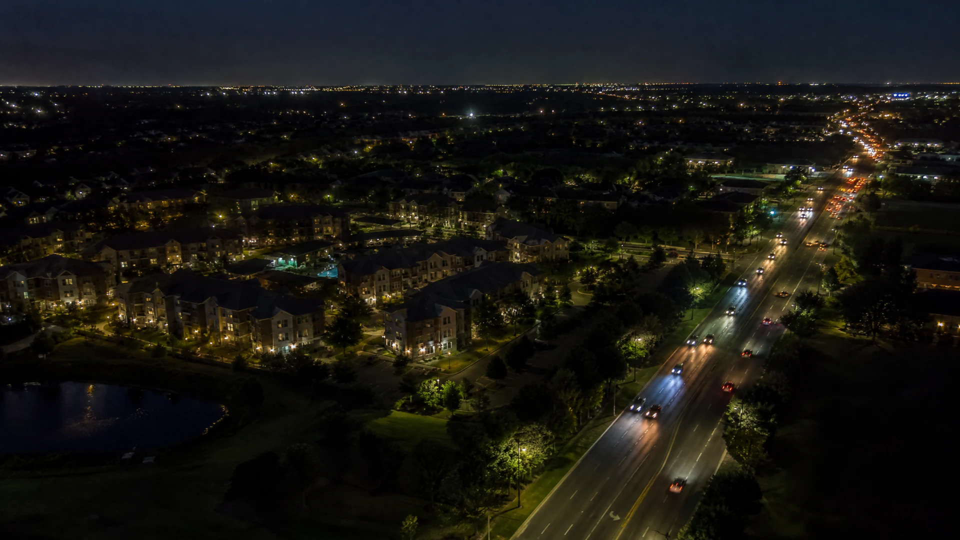 Aerial night view of a suburban road with moving vehicles and illuminated apartment complexes surrounded by trees.