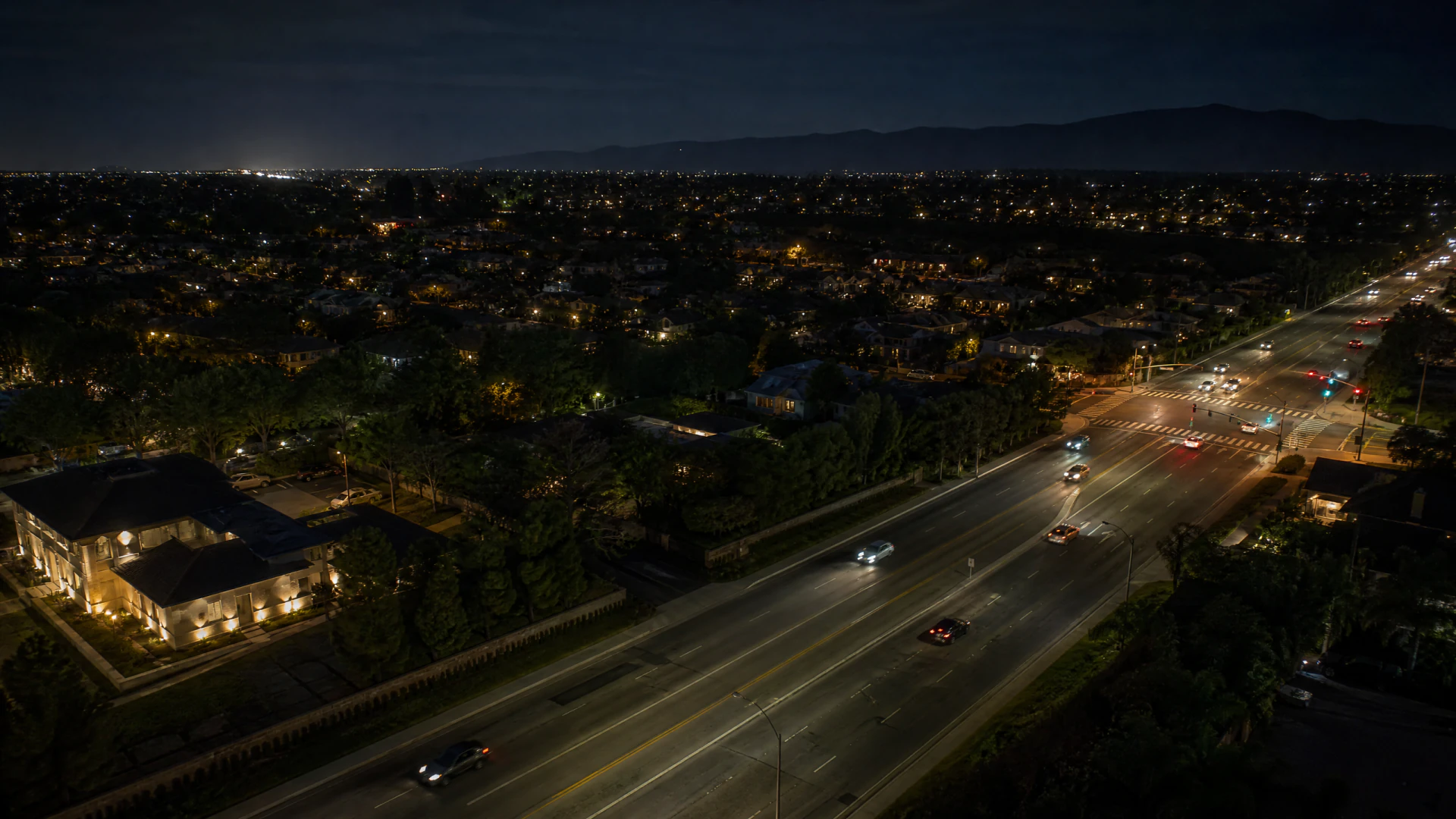 Aerial night photograph of a multi-lane urban boulevard lined with streetlights, with cars in motion and a sprawling lit cityscape in the background.
