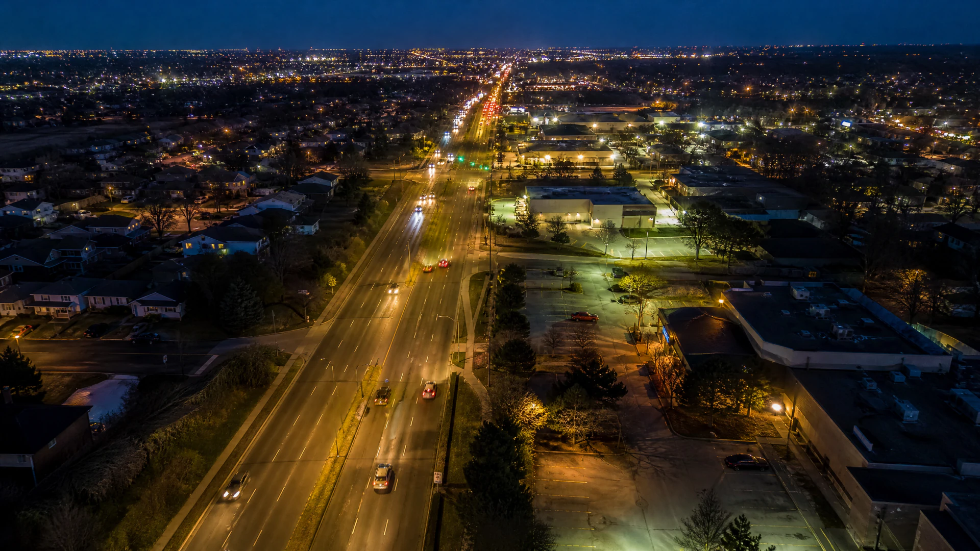 Aerial nighttime view of a wide suburban arterial road lined with commercial buildings, parking lots, and residential neighborhoods illuminated by streetlights.