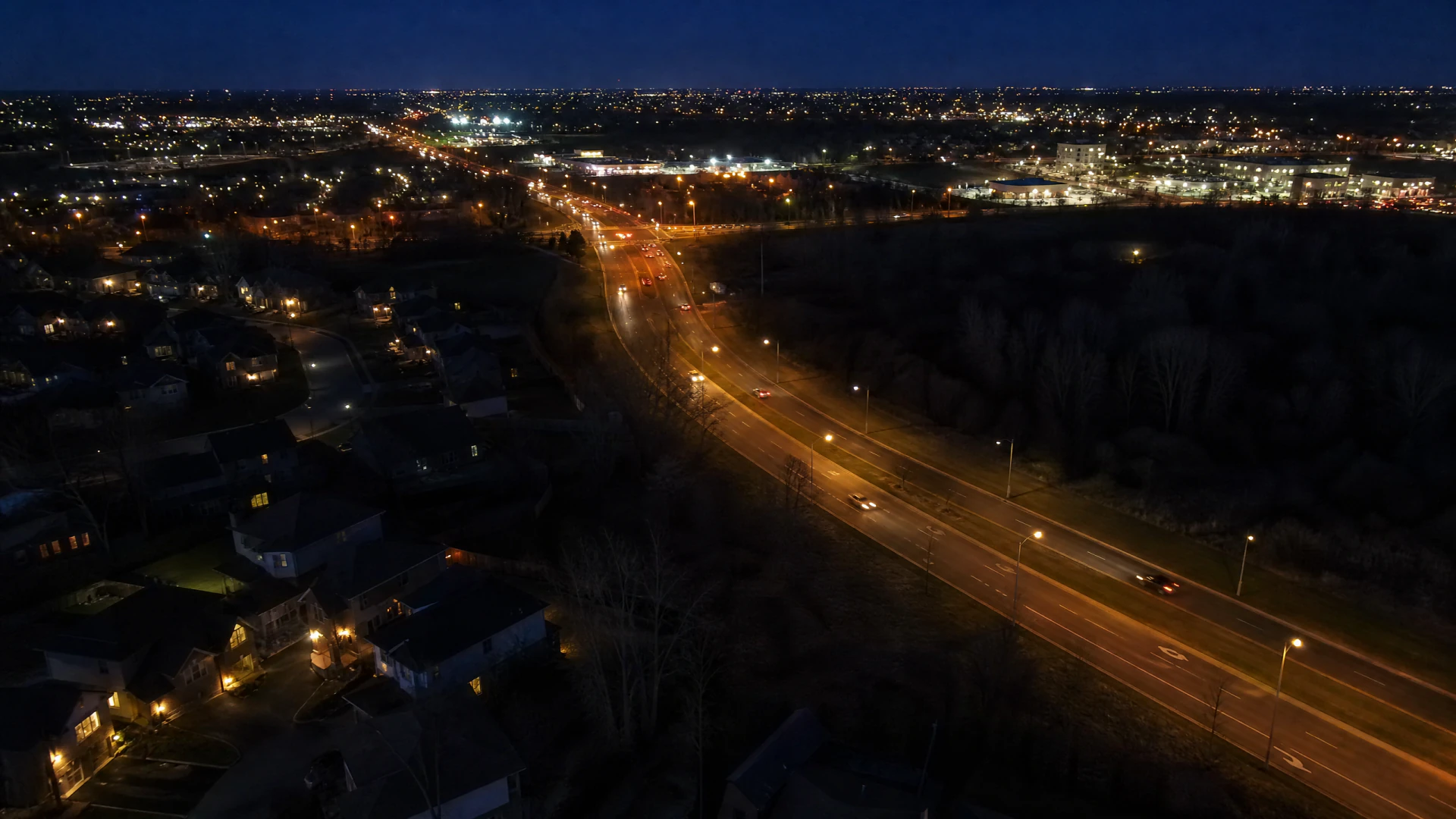 Aerial night view of a suburban multi-lane road lined with orange streetlights, flanked by residential housing on the left and bare trees on the right, leading toward a distant commercial district.