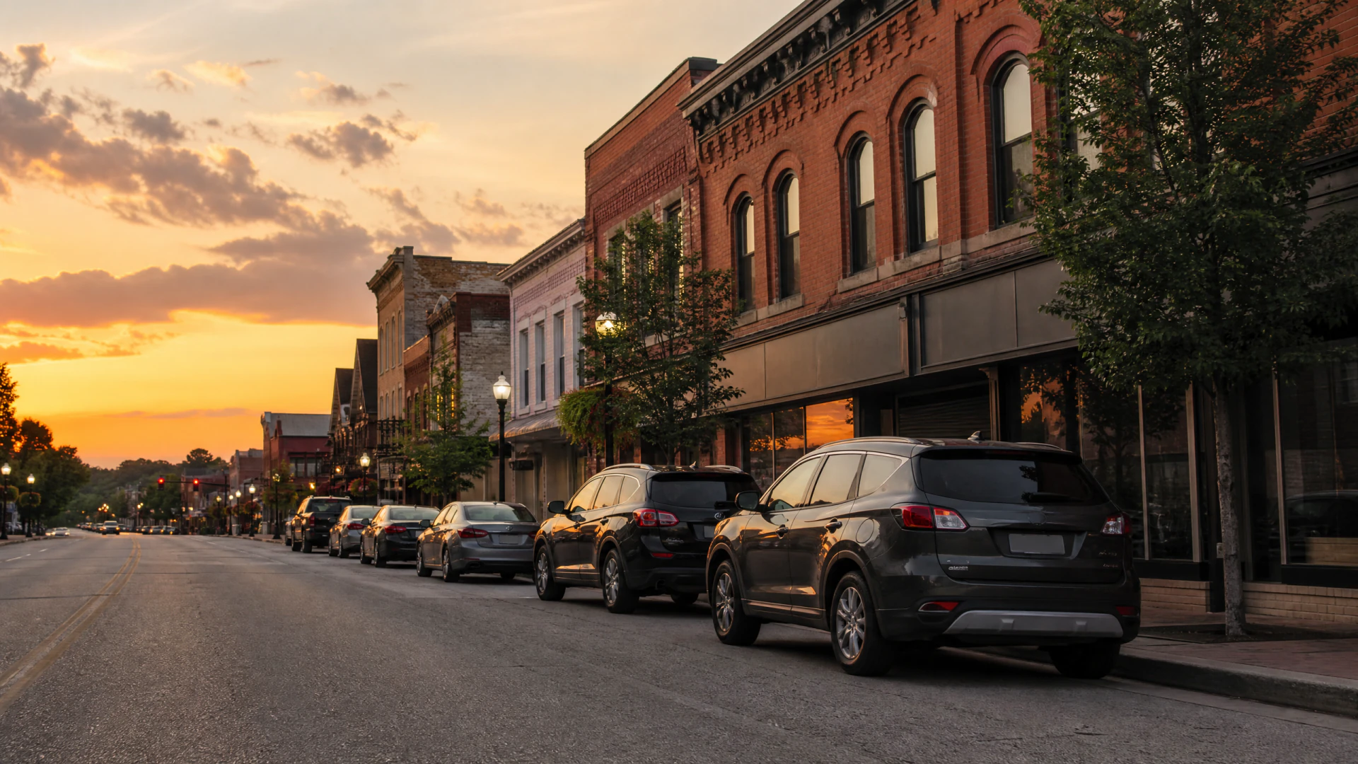 Parked cars line a historic brick-building main street under a golden sunset sky.