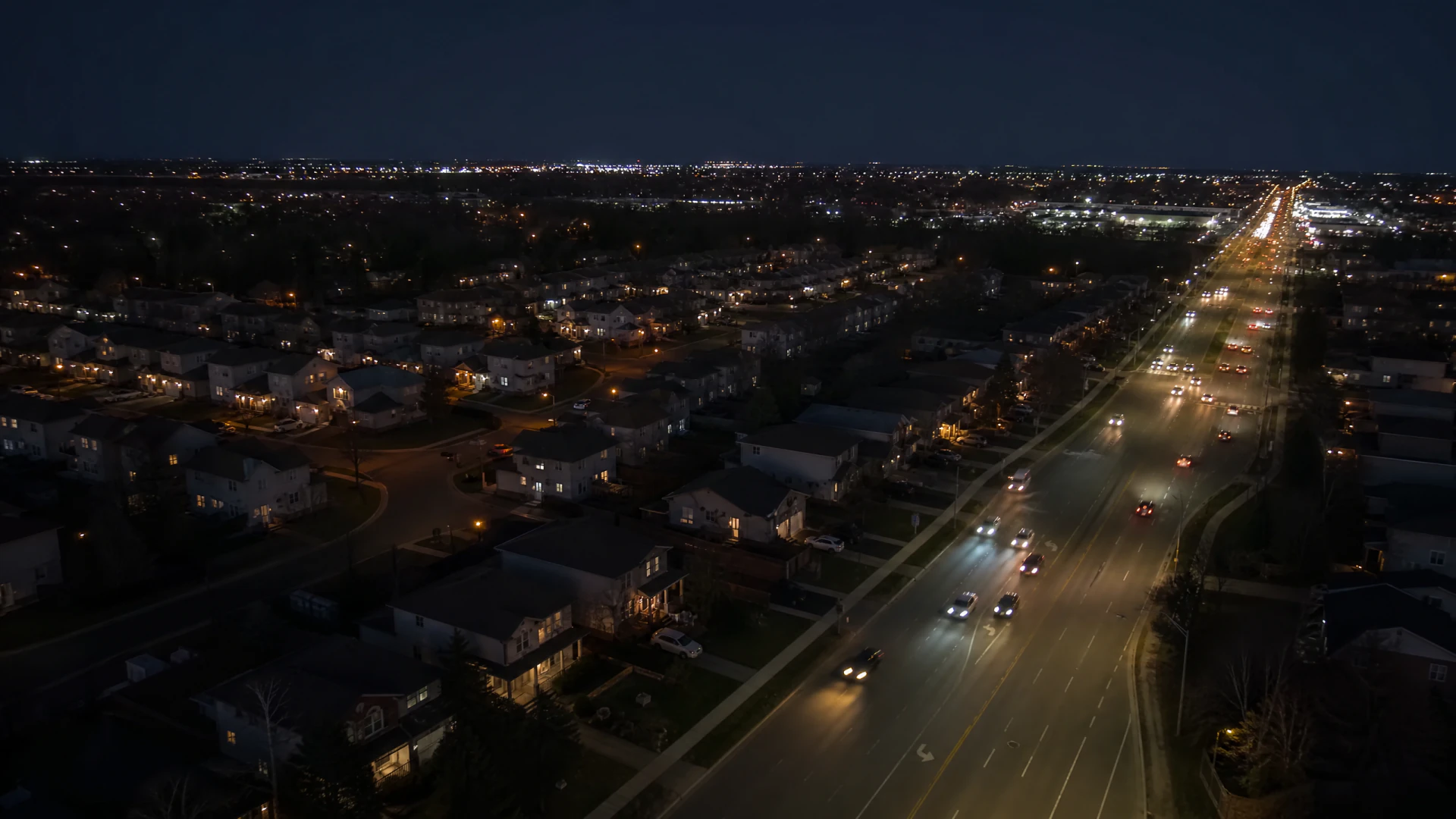 Aerial nighttime photograph of a suburban neighborhood alongside a multi-lane road with moving vehicles and streetlights.